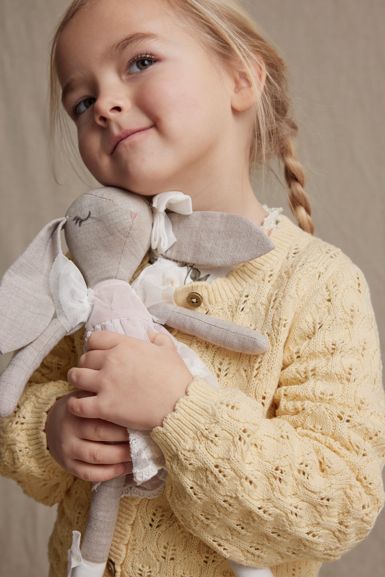 Close-up front view of a kid in a yellow pattern-knitted cardigan, holding a grey bunny doll.
