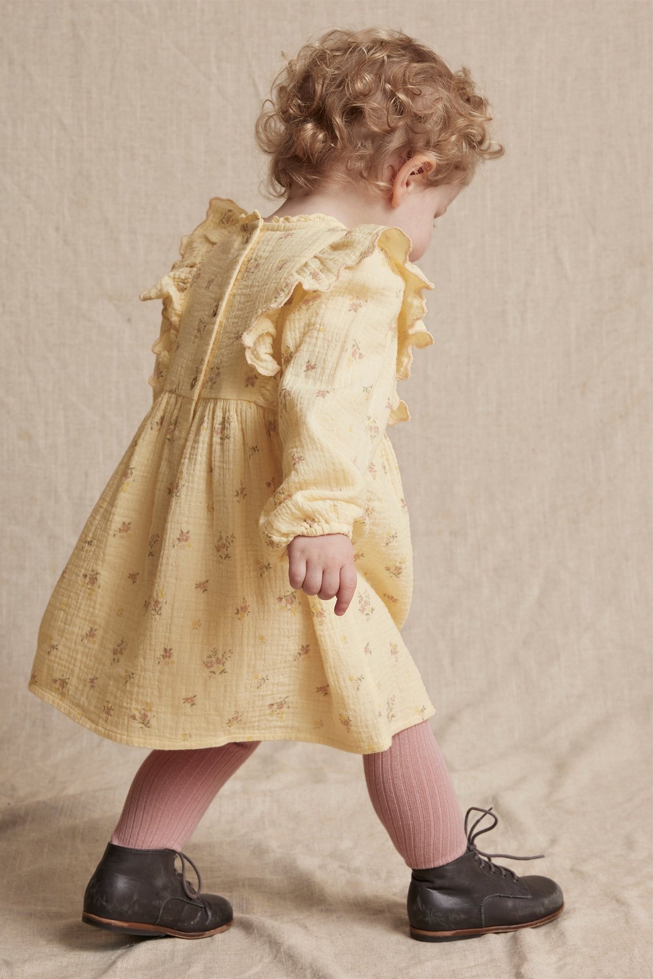 Back view of a baby wearing a yellow floral crumpled cotton dress with ruffles, pink tights, and dark grey boots.