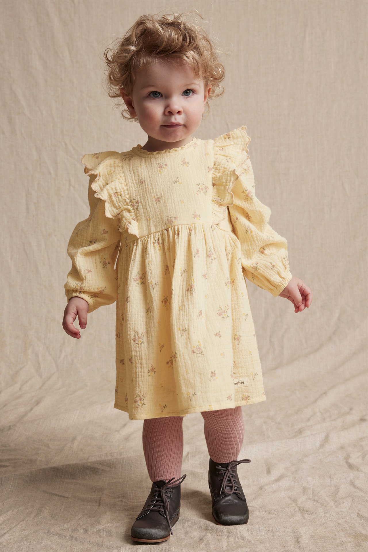 Front view of a baby in a yellow floral organic cotton dress with ruffles, pink tights, and dark brown boots.