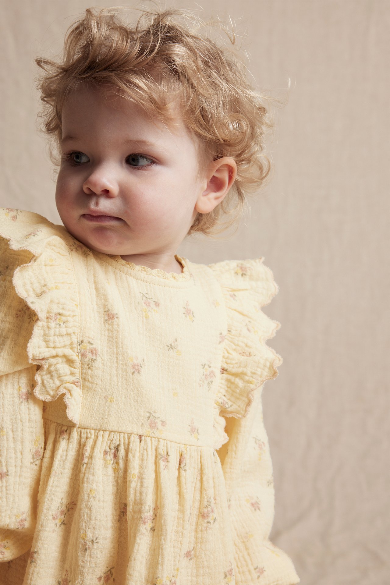 Close-up of a baby wearing a light yellow floral cotton dress with ruffles and lace trim.