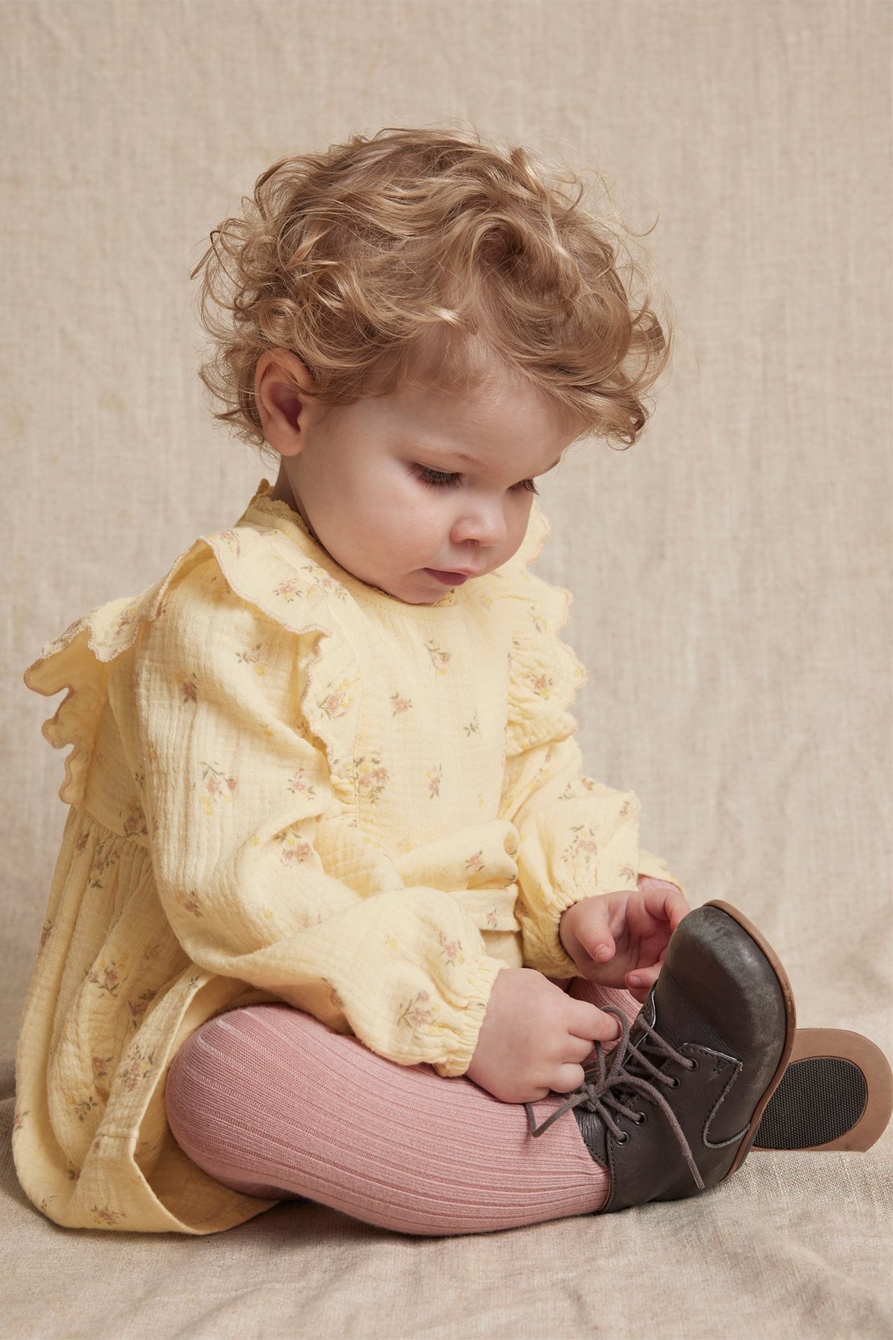 Front view of a baby in a yellow floral cotton dress, pink ribbed tights, and dark grey lace-up shoes.