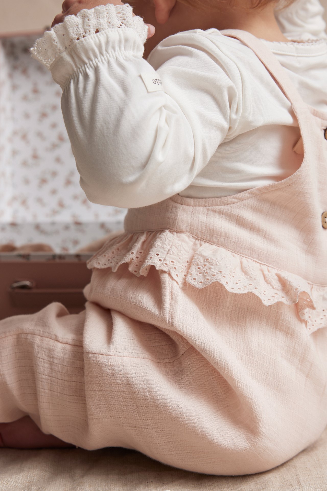 Seitenansicht eines Babys in hellrosa Baumwoll-Latzhose mit Spitzenrüschen über einem weißen Langarmshirt.