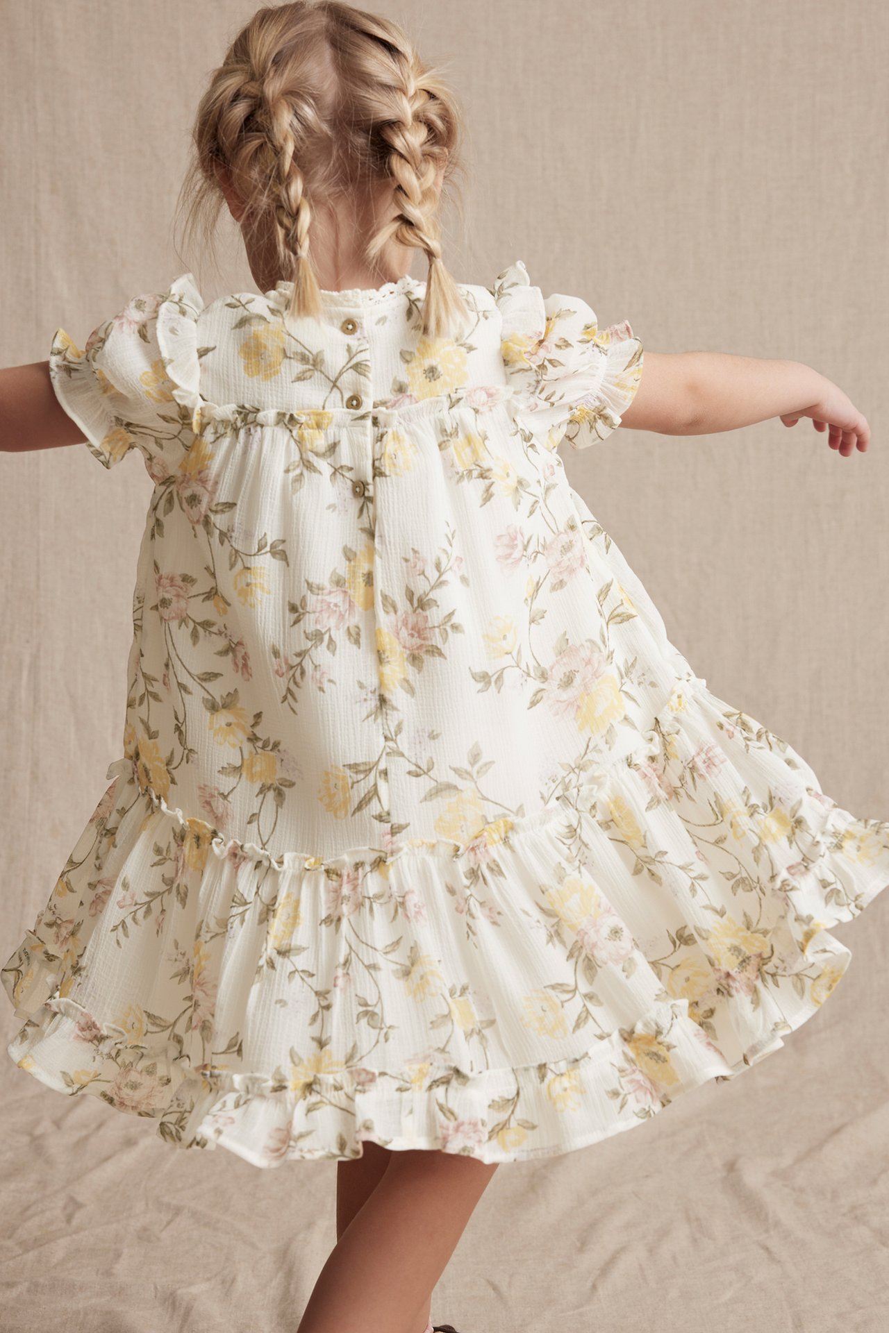Rückenansicht eines Kindes in einem cremefarbenen geblümten Chiffonkleid mit Rüschenärmeln, Stufenrock und Knöpfen hinten.
