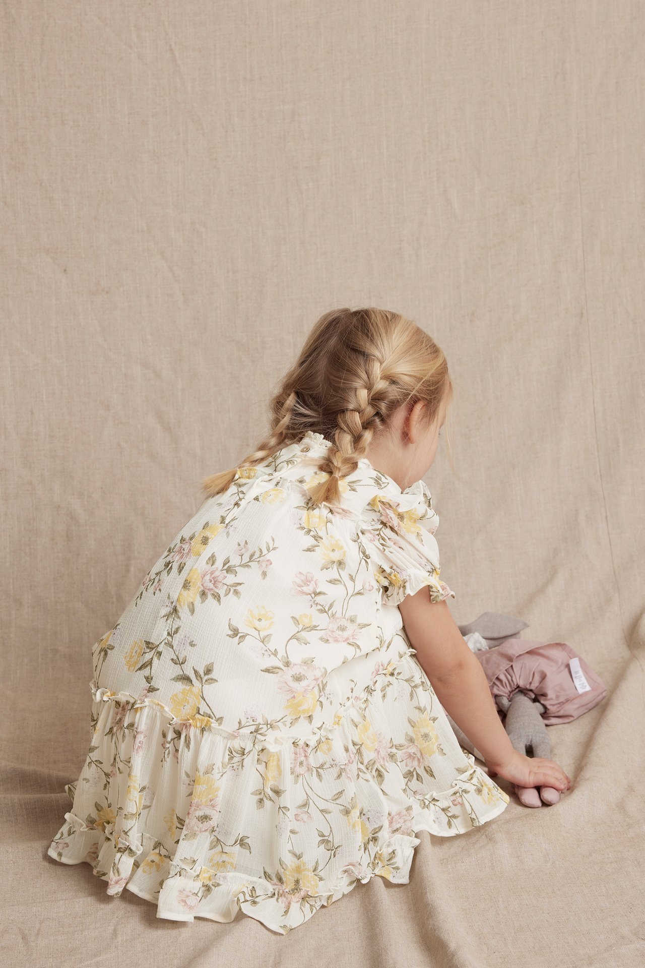 Rückenansicht eines Kindes in einem cremefarbenen geblümten Chiffonkleid mit Rüschen, neben einem grauen Spielzeug.