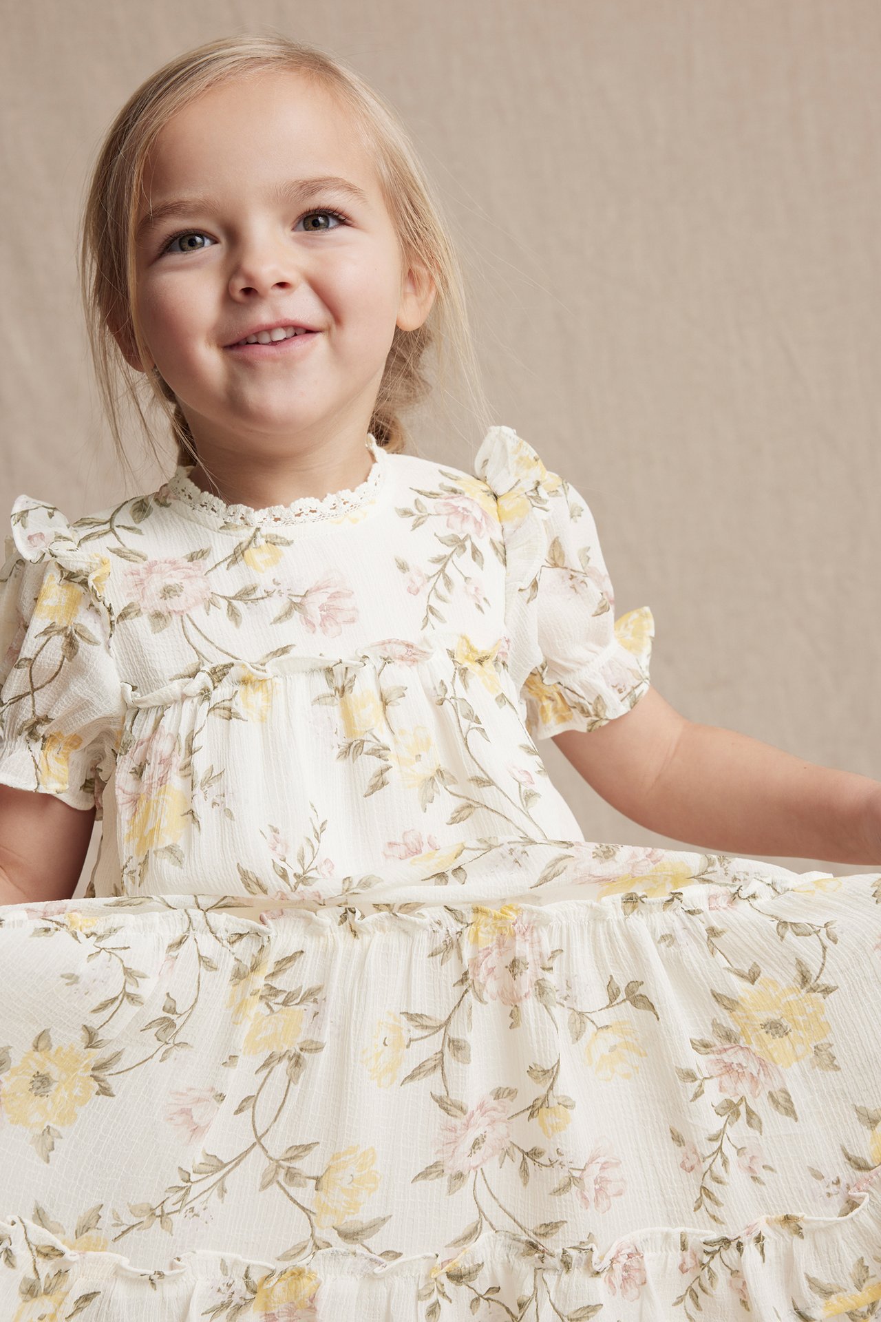 Vorderansicht eines lächelnden Kindes in einem cremefarbenen Blumen-Chiffonkleid mit Spitze und Puffärmeln.