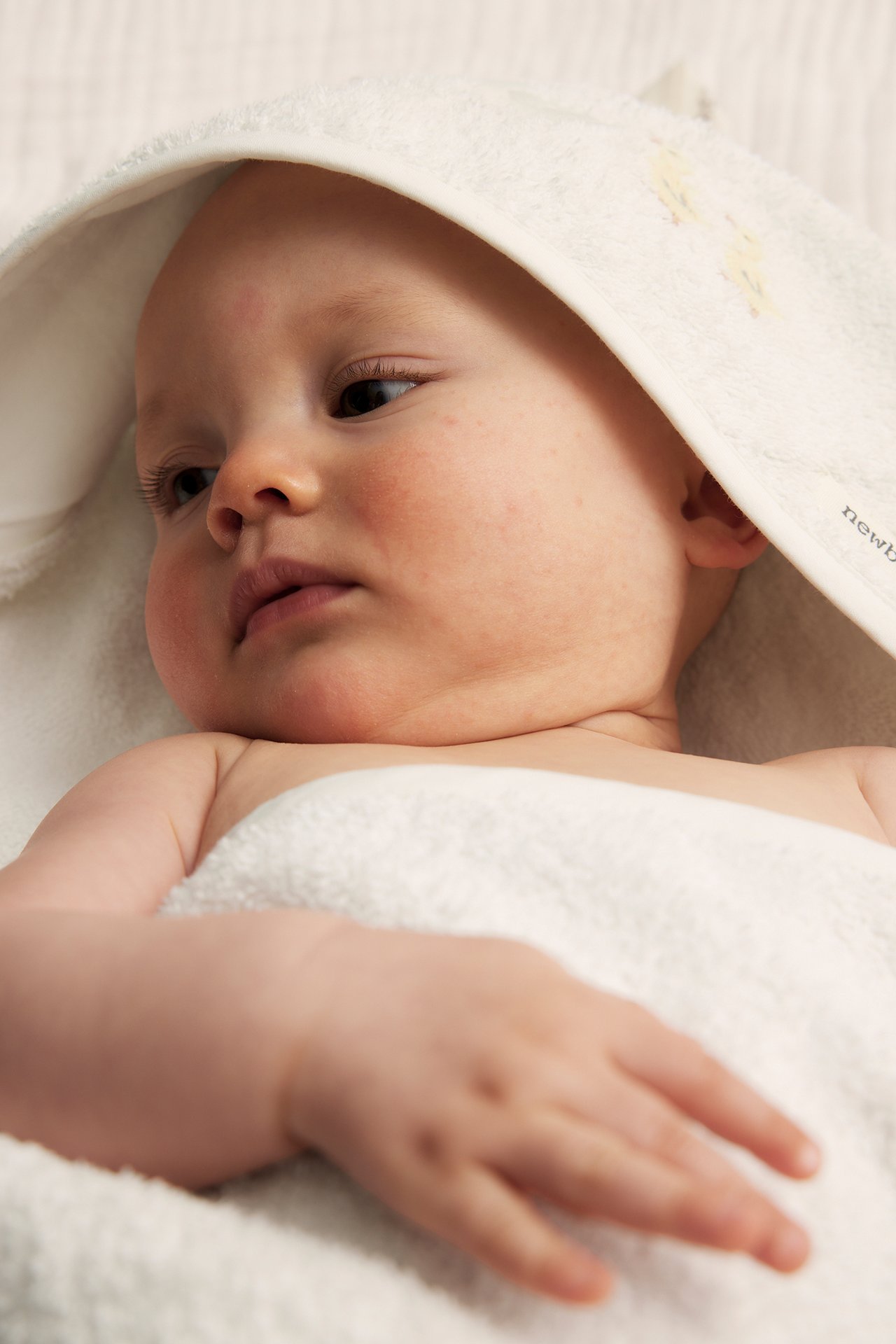 Nahaufnahme eines Babys, eingewickelt in ein cremefarbenes Kapuzen-Frotteehandtuch mit Stickerei.