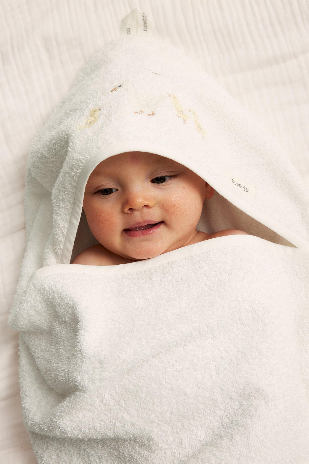 Nahaufnahme eines lächelnden Babys, eingewickelt in ein cremefarbenes Kapuzen-Frotteehandtuch mit Entenstickerei.