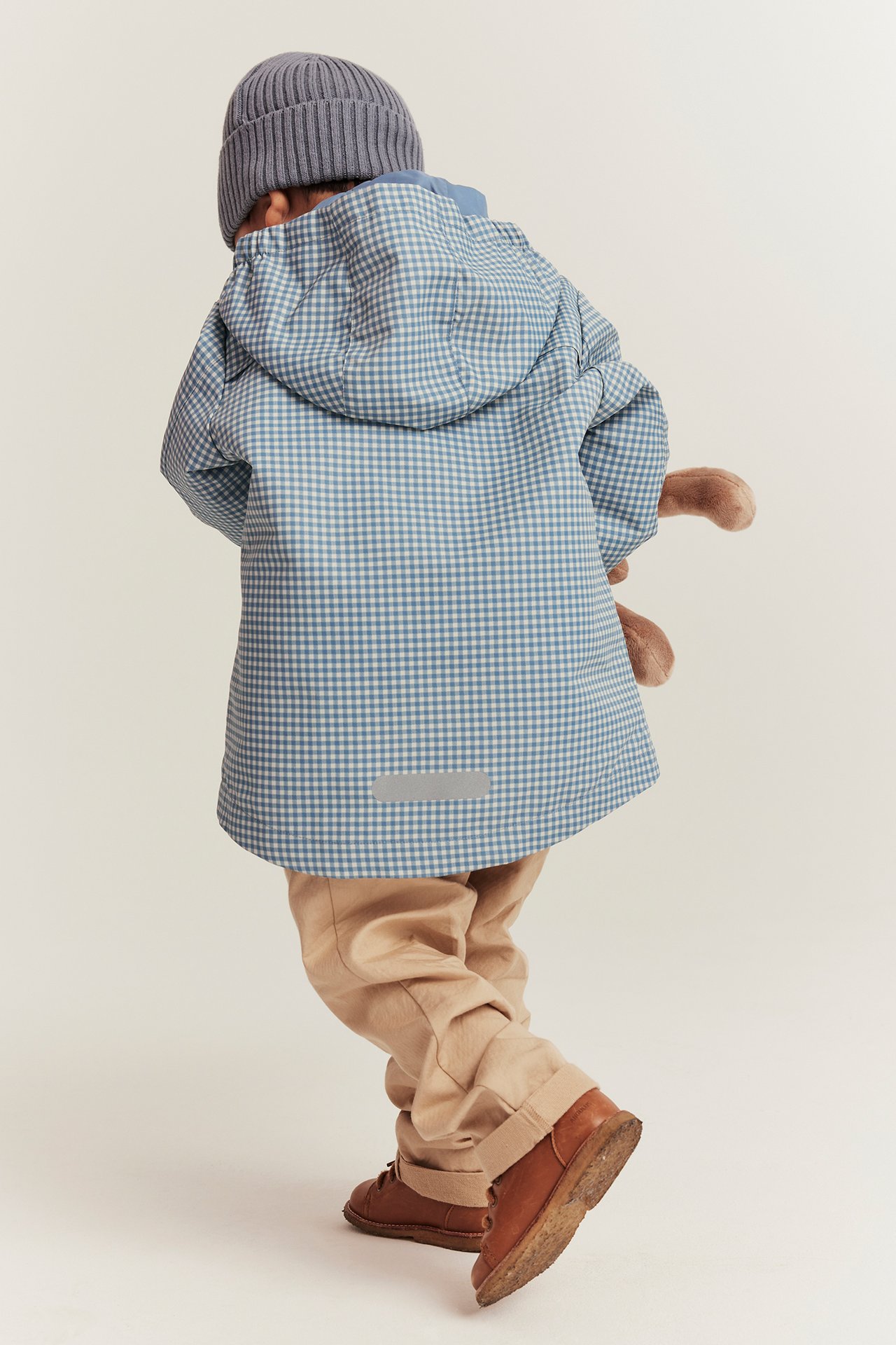 Rückenansicht: Kind in blau karierter Jacke, grauer Mütze, beiger Hose, braunen Schuhen, hält ein Plüschtier.