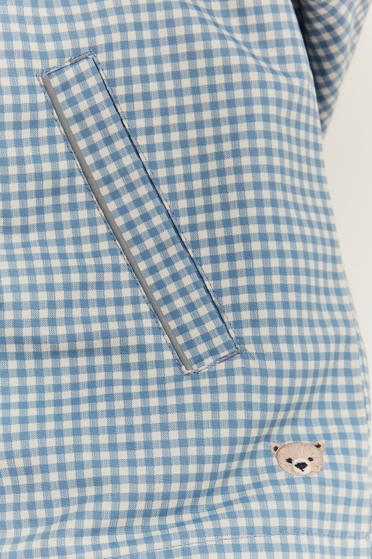 Nahaufnahme einer blau-weiß karierten Kinderjacke mit reflektierender Tasche und Teddybär-Stickerei.