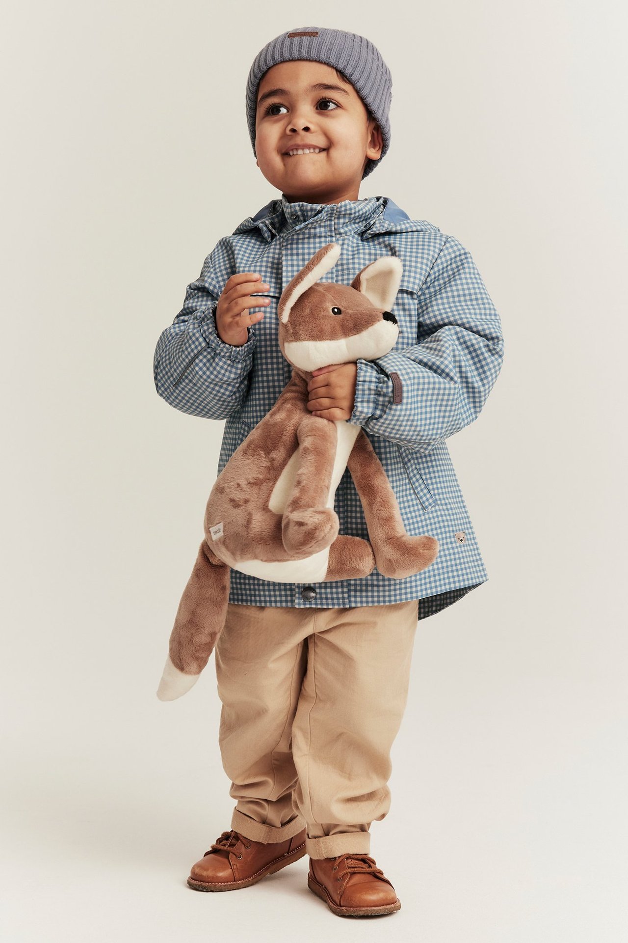 Vorderansicht: Kind in blau karierter Kapuzenjacke, beiger Hose, braunen Schuhen, grauer Mütze, hält Fuchsplüschtier.