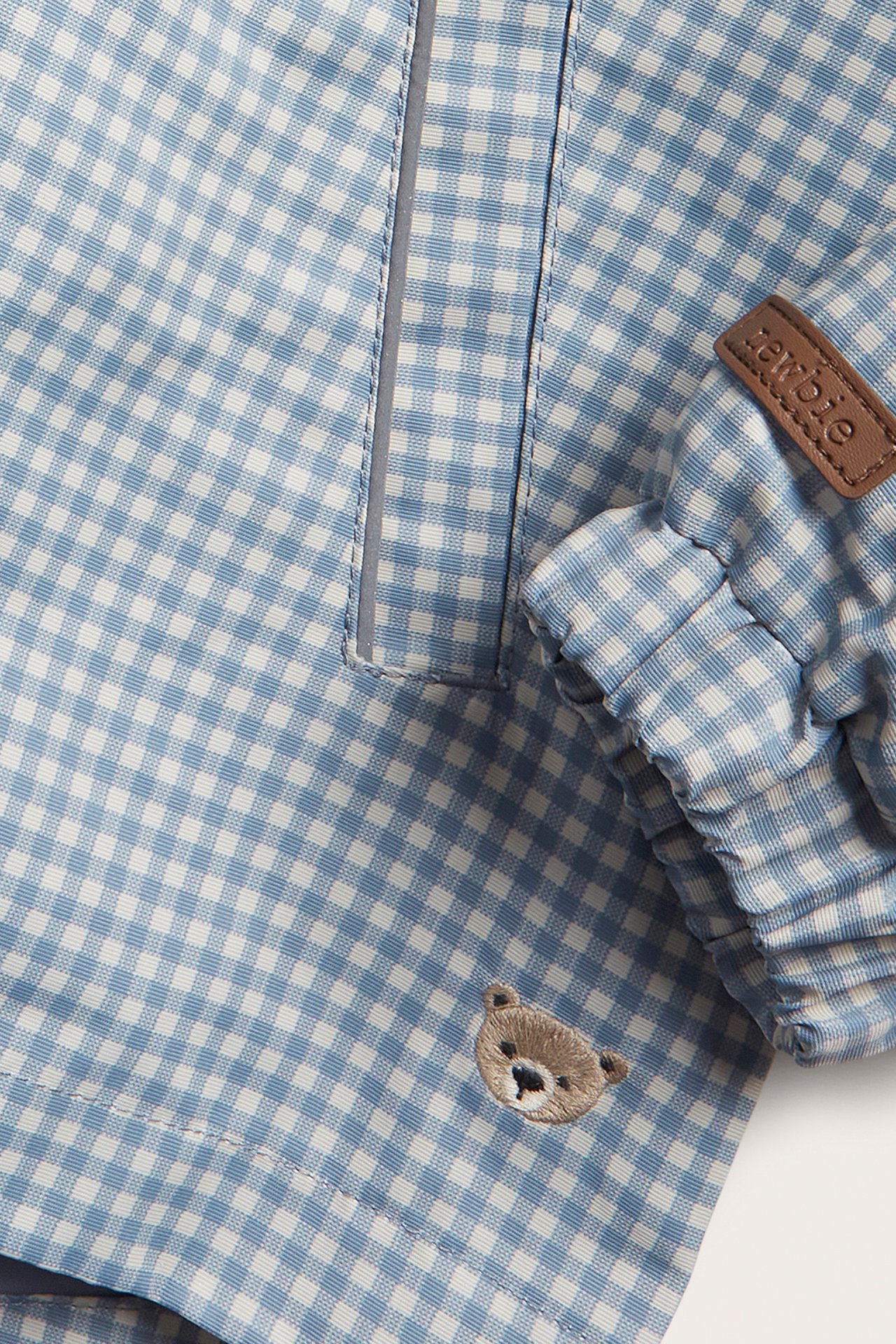 Nahaufnahme einer blau-weiß karierten Kinderjacke mit elastischem Bündchen, Reflexstreifen und gesticktem Bären.