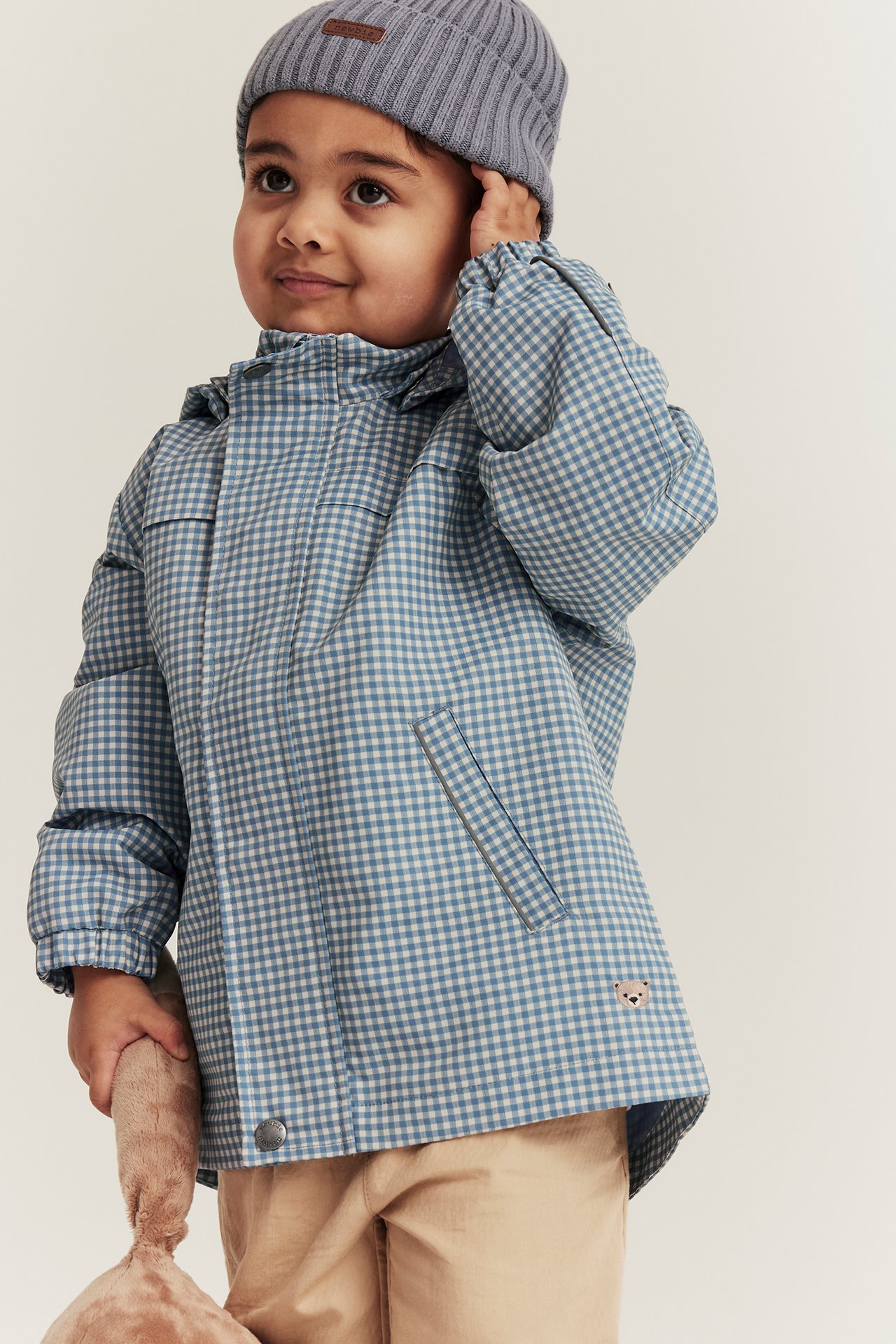 Vorderansicht: Kind in blau karierter Kapuzenjacke, beiger Hose, grauer Mütze, hält ein braunes Plüschtier.
