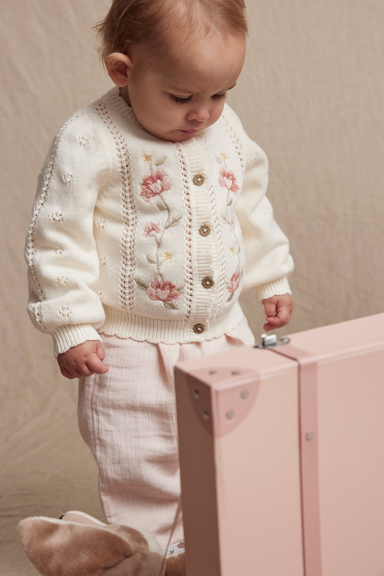 Front view of a baby in an off-white organic cotton cardigan with floral embroidery, pink trousers, and a pink suitcase.