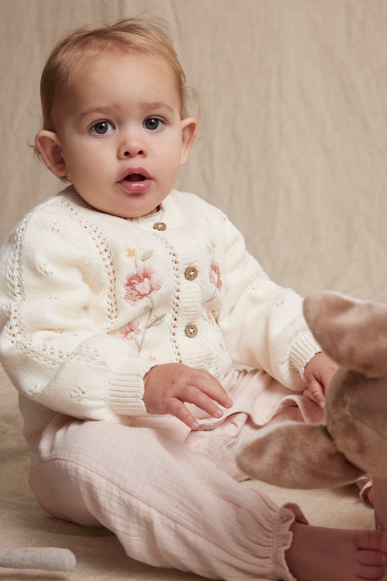 Front view: baby in an off-white cotton cardigan with floral embroidery, pointelle knit, and light pink trousers.