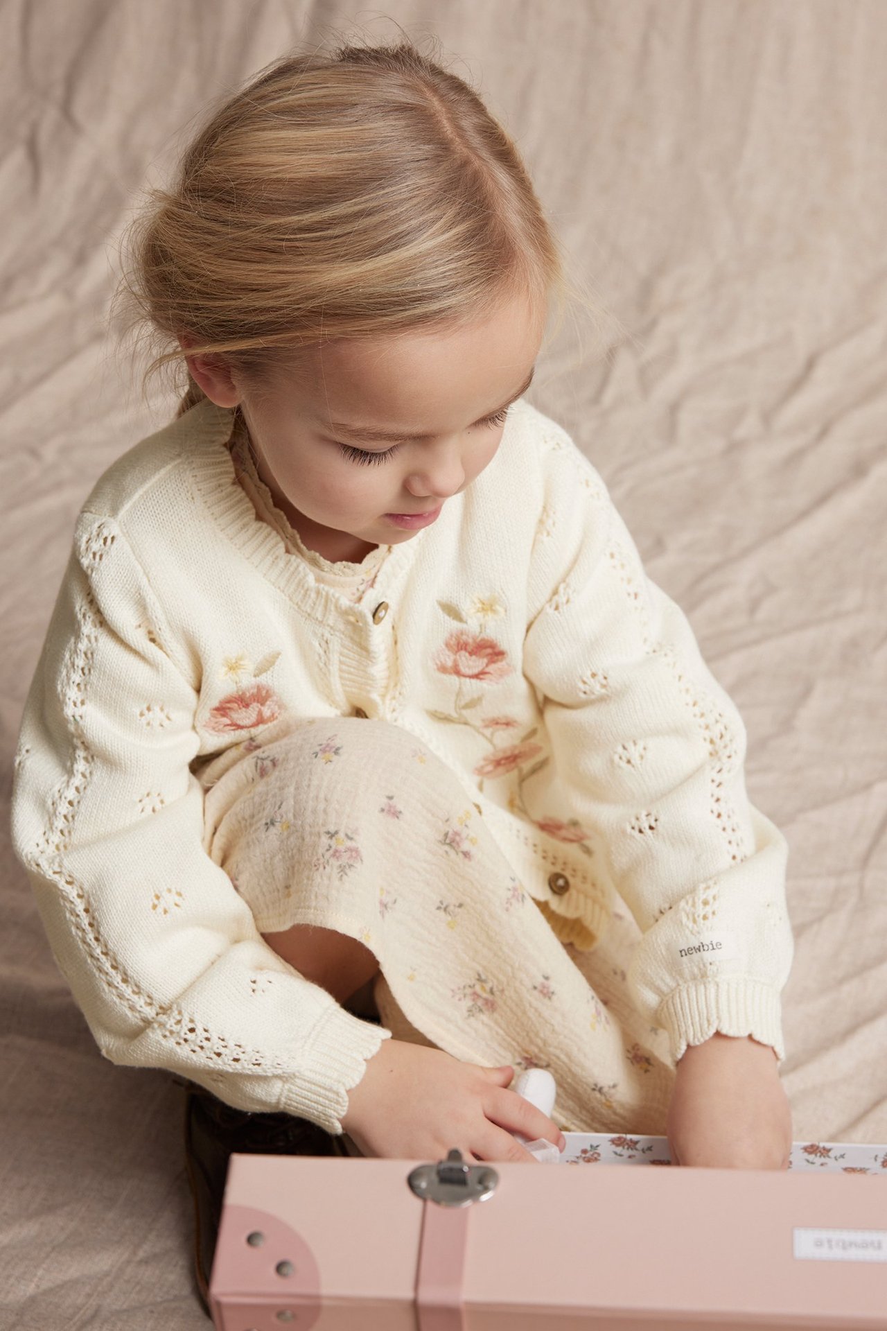 Front view of a kid in an offwhite cotton cardigan with floral embroidery, pointelle pattern, and light floral leggings.