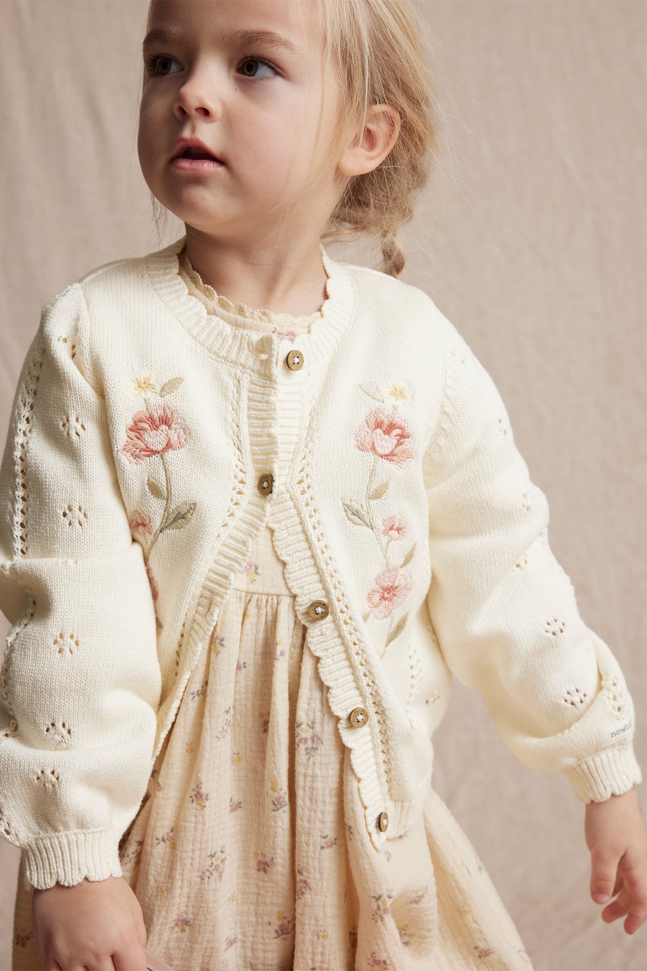 Front view: Kid girl in offwhite cotton cardigan with floral embroidery over a light floral dress.