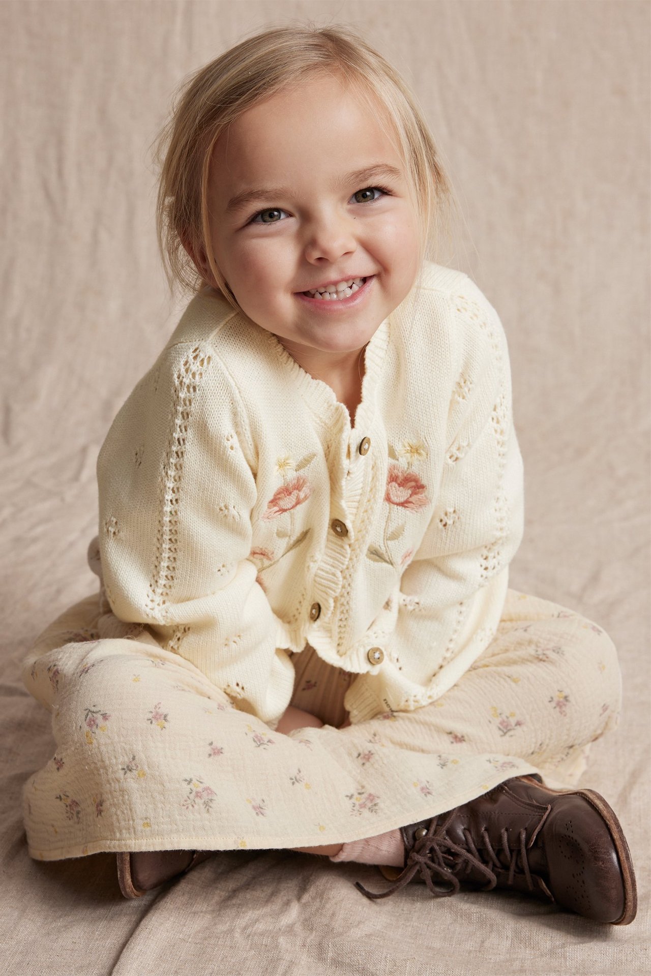 Front view of a smiling kid wearing an off-white cotton cardigan with floral embroidery, light floral pants, and brown boots.