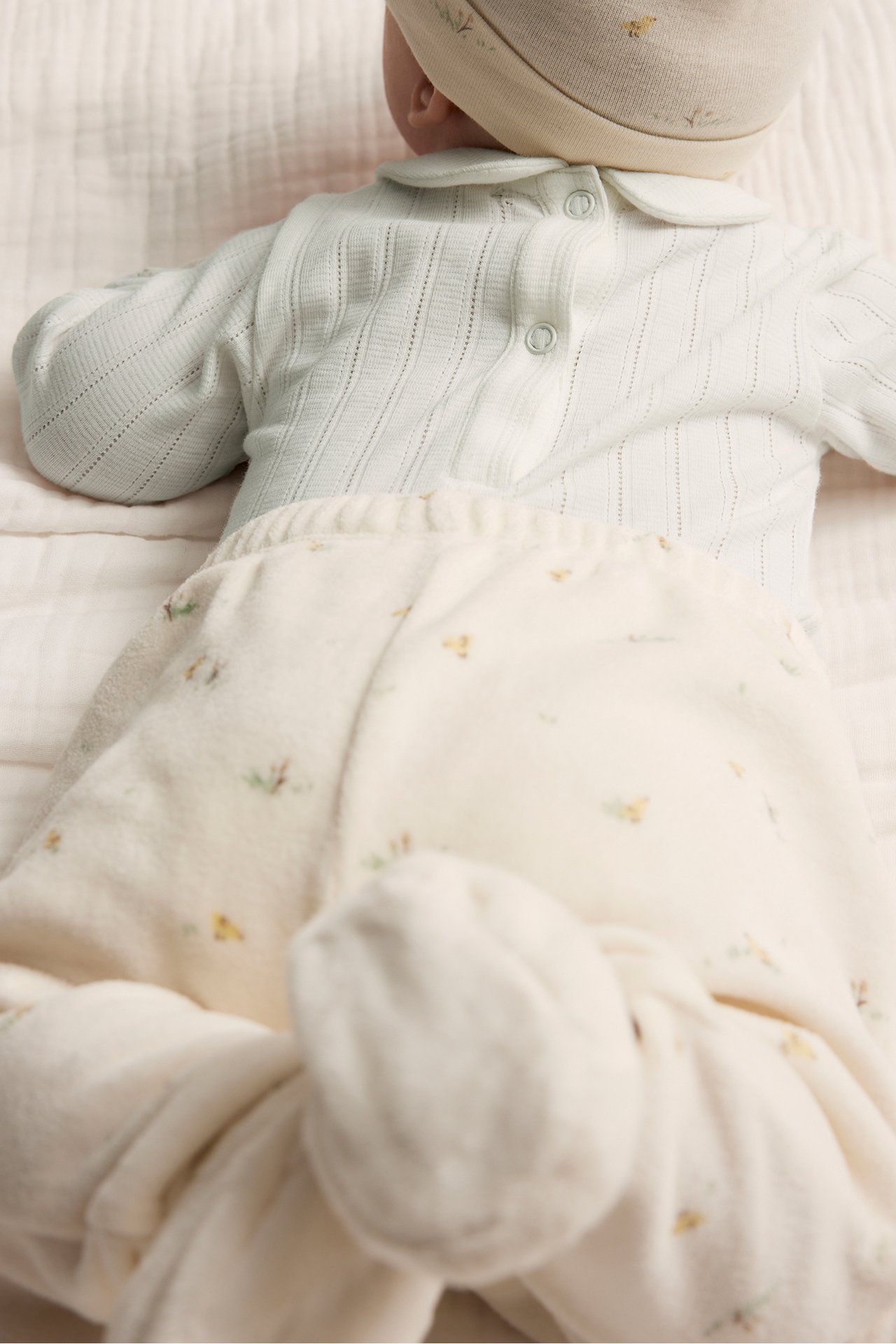 Back view of a baby in an off-white pointelle rib collared bodysuit, floral pants, and a beige hat.