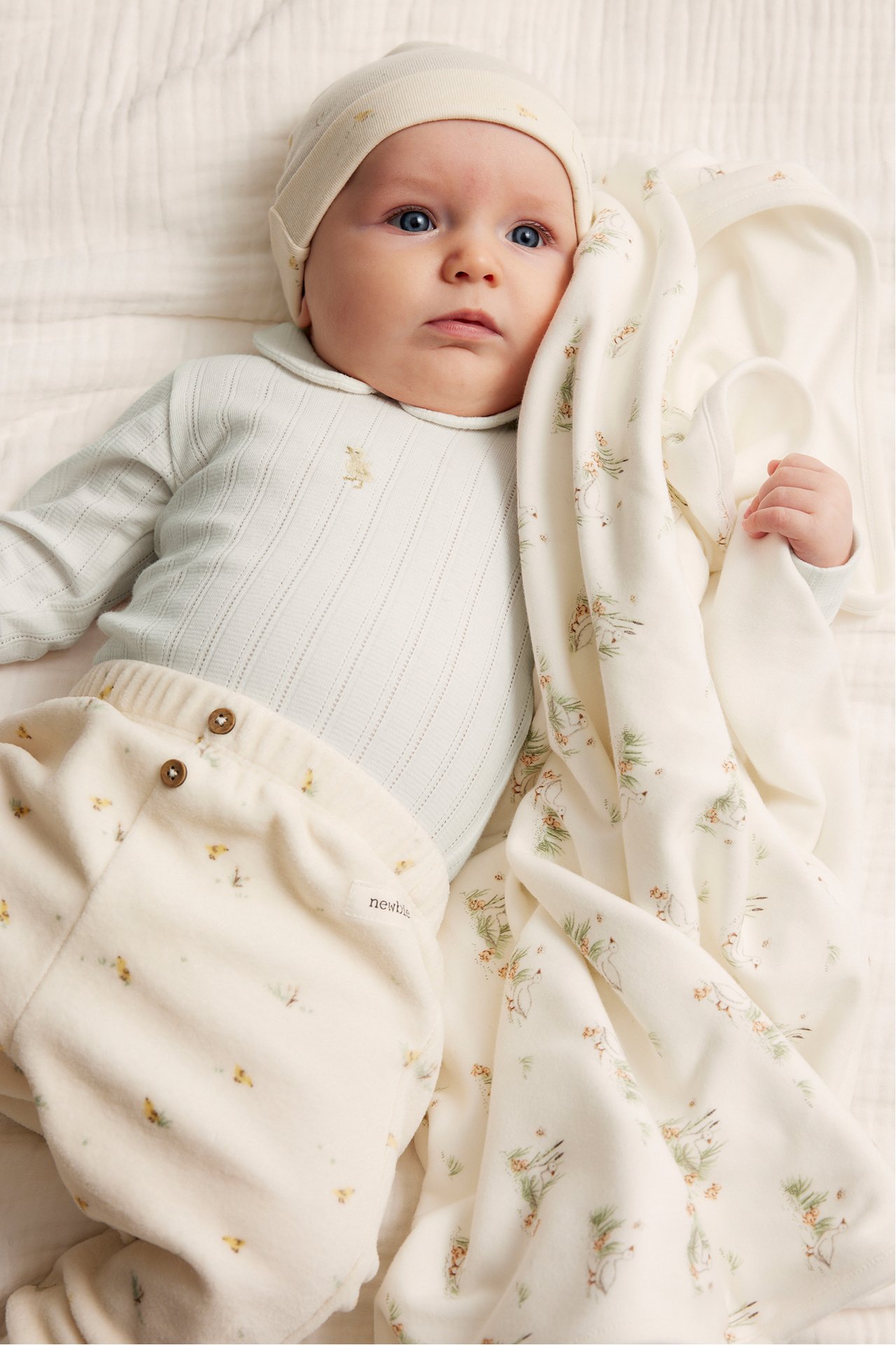 Baby in an offwhite ribbed body with collar, offwhite patterned pants, beanie, and a patterned blanket.