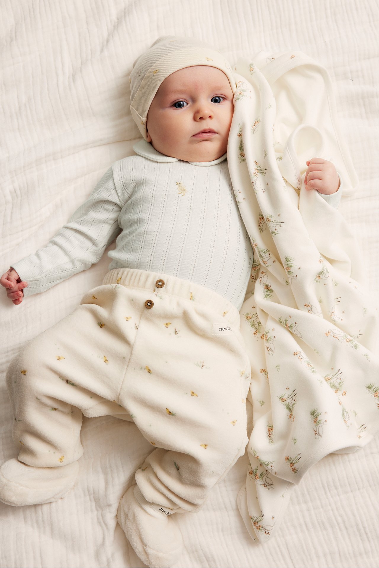 Front view of a baby in an off-white pointelle rib body with collar, patterned pants, hat, and blanket.