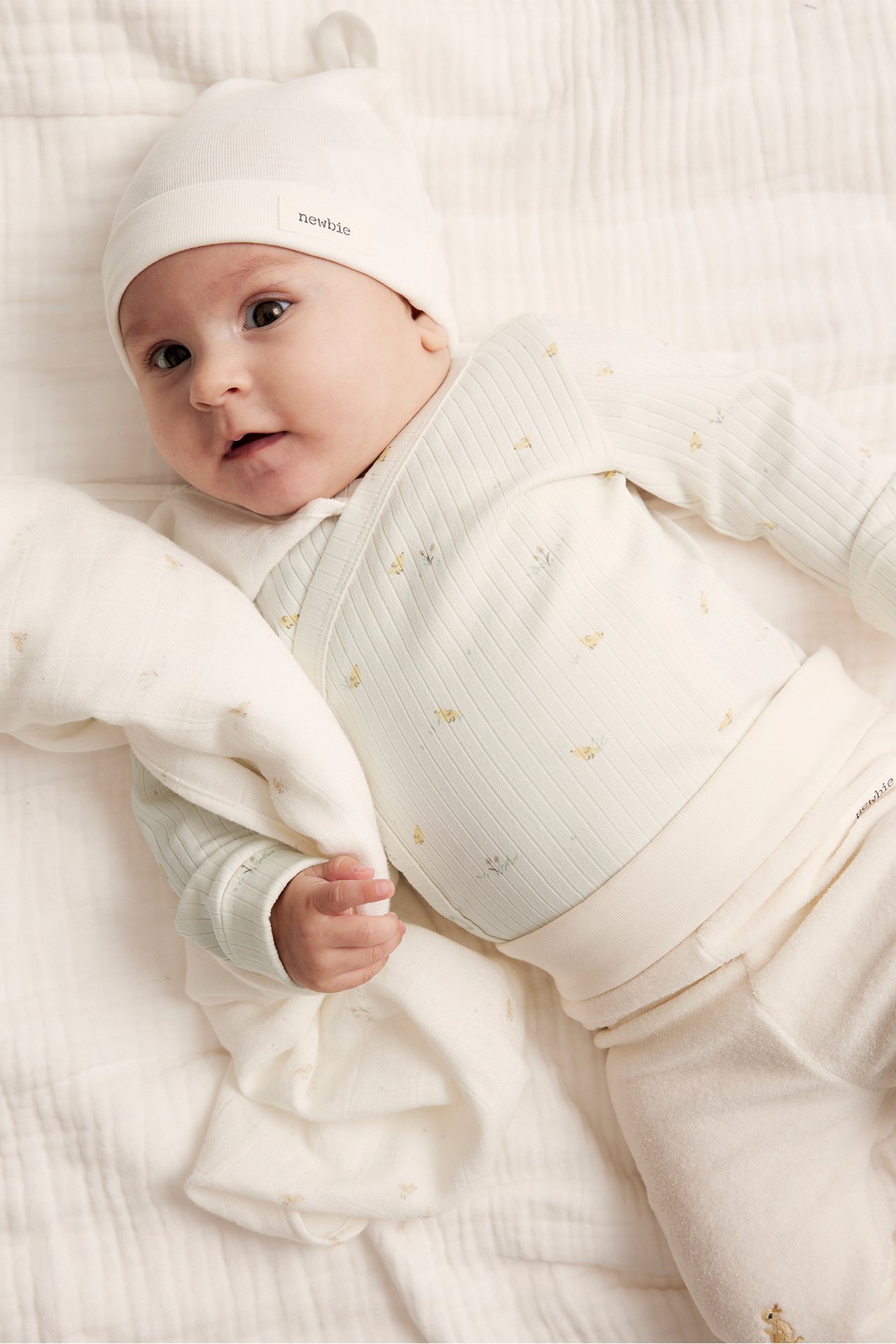 Top view of baby in off-white ribbed wrap bodysuit with floral print, off-white hat, trousers, holding blanket.