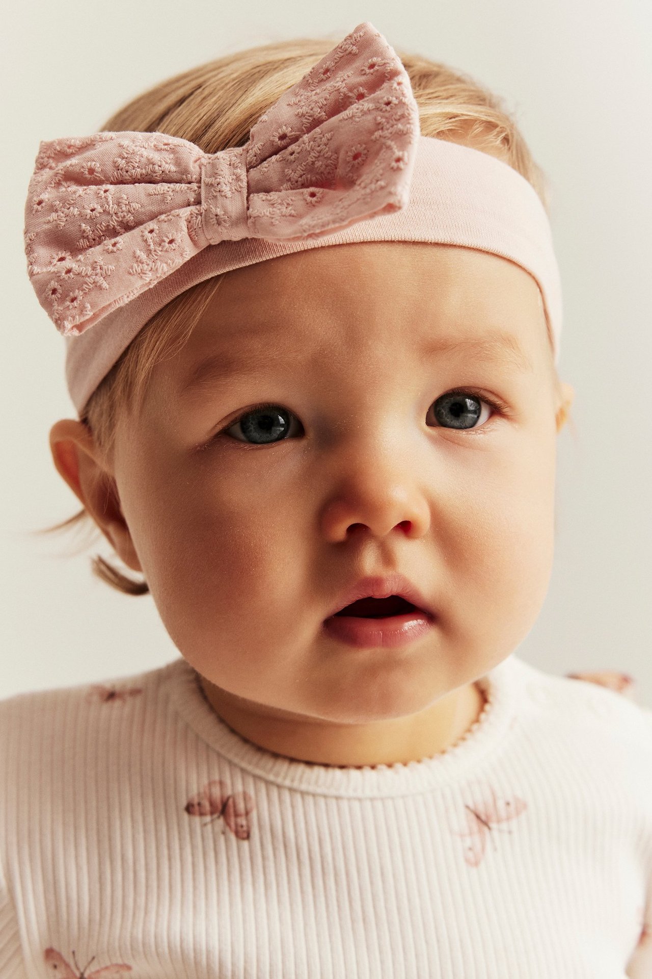Close-up of a baby wearing a light pink jersey hairband with a lace bow and a ribbed butterfly print top.
