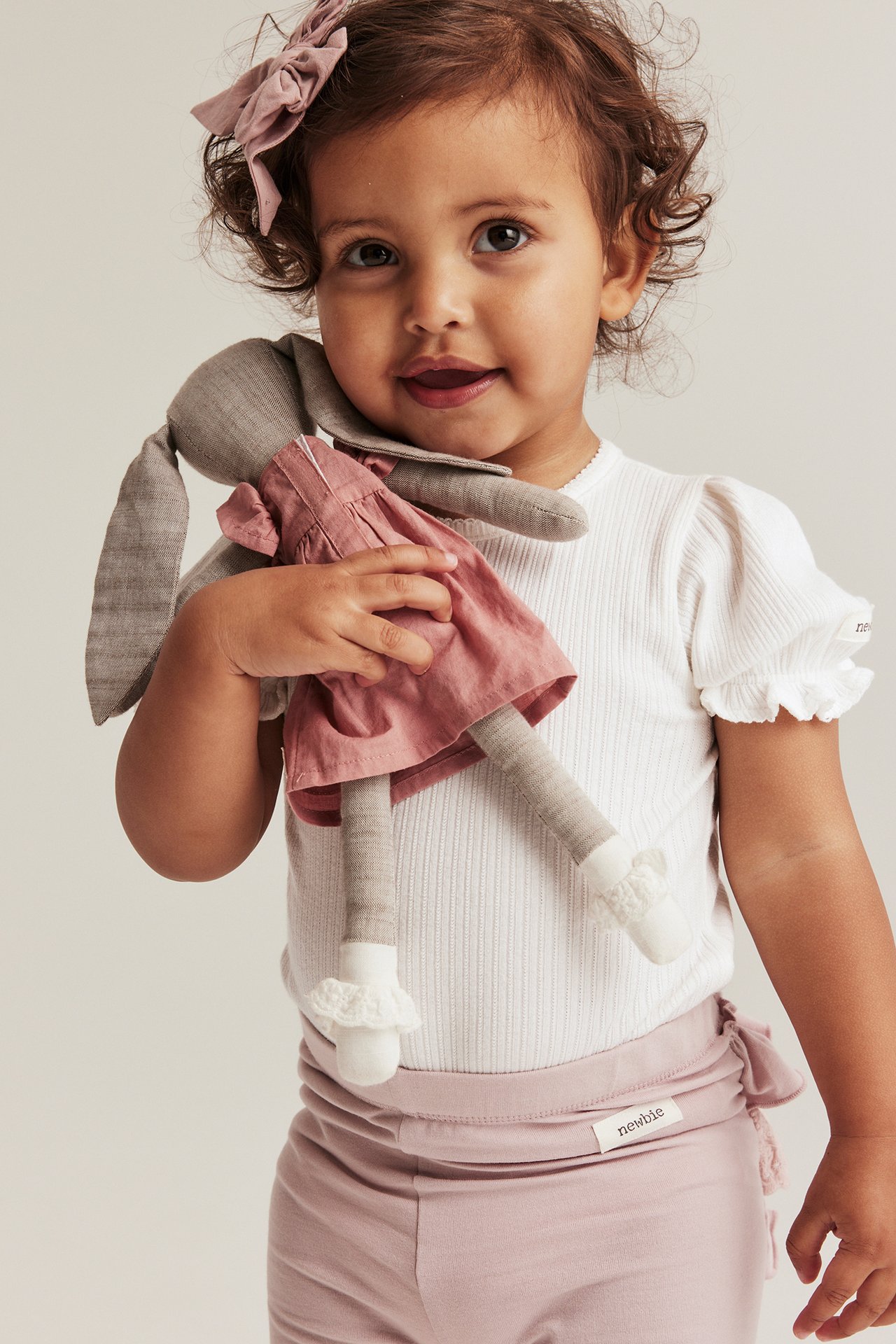 Front view: baby in off-white ribbed puff-sleeved bodysuit, pink leggings, bow, holding bunny doll.