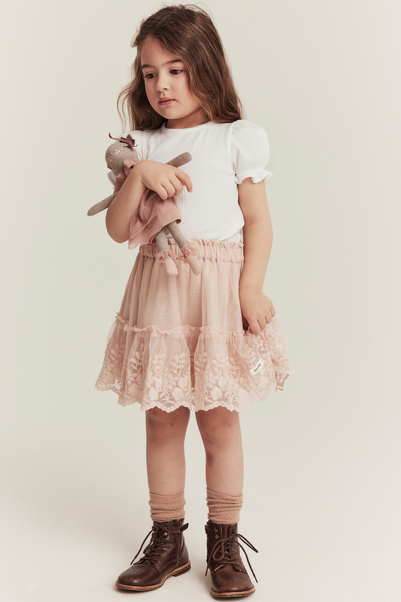 Vorderansicht: Kind in geripptem Off-White Puffärmel-Top, rosa Spitzrock, braunen Stiefeln, hält eine Puppe.