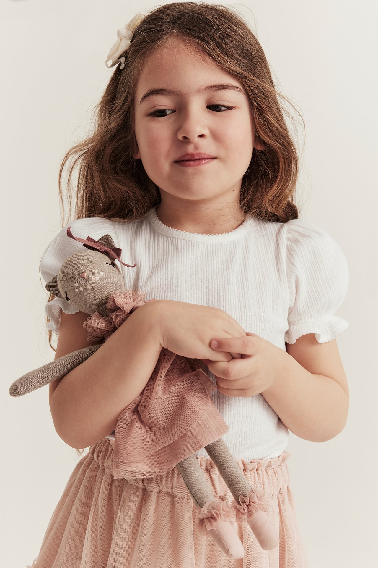 Vorderansicht: Kind in cremeweißem geripptem Puffärmel-Top und rosa Tüllrock, hält eine Stoffpuppe.