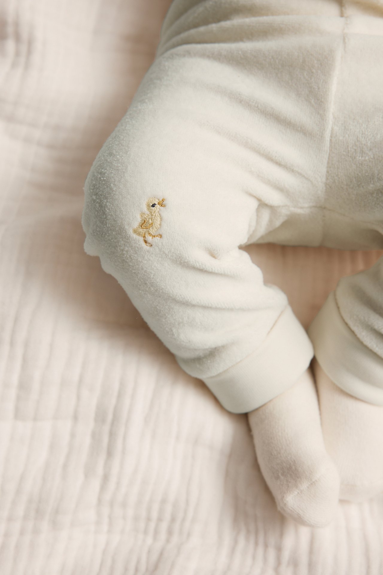 Close-up of baby's legs in off-white velour leggings with a chick embroidery and foldable cuffs, plus white socks.