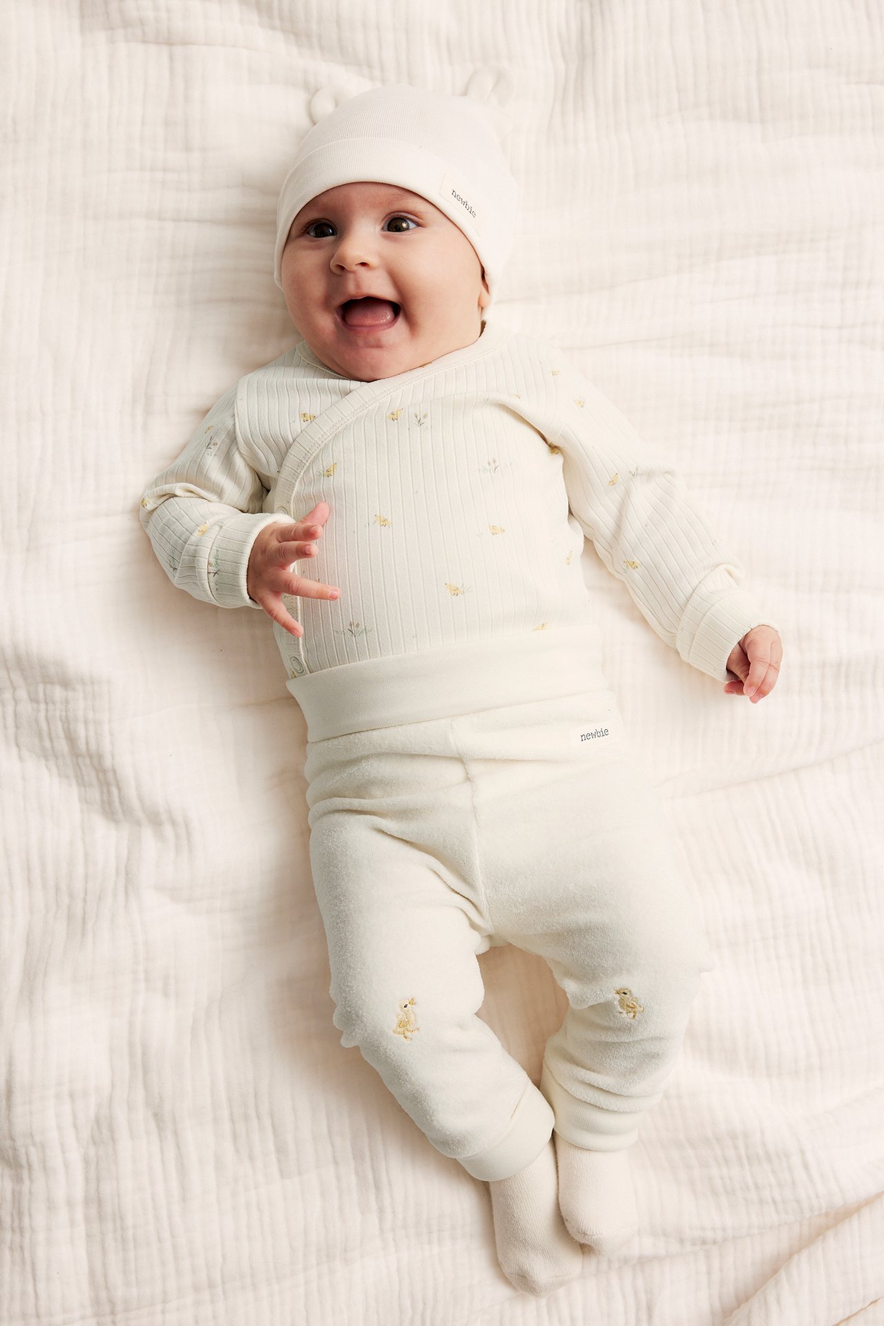 Top-down view of a baby in offwhite velour leggings with duck embroidery, a ribbed wrap top, and a hat.