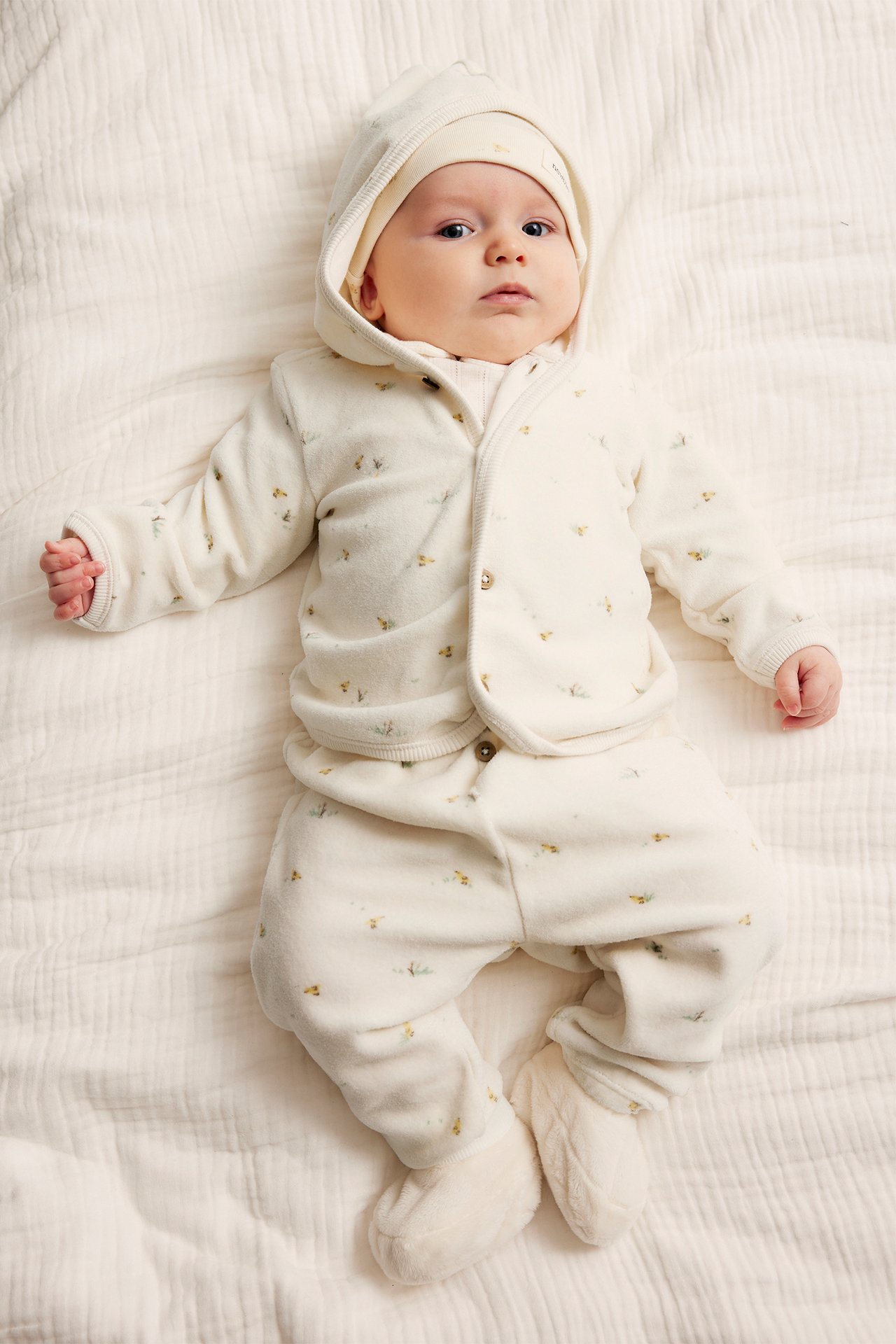 Draufsicht: Baby in einem cremefarbenen gemusterten Velours-Kapuzenpullover mit Knöpfen, passender Hose, Mütze und Schühchen.