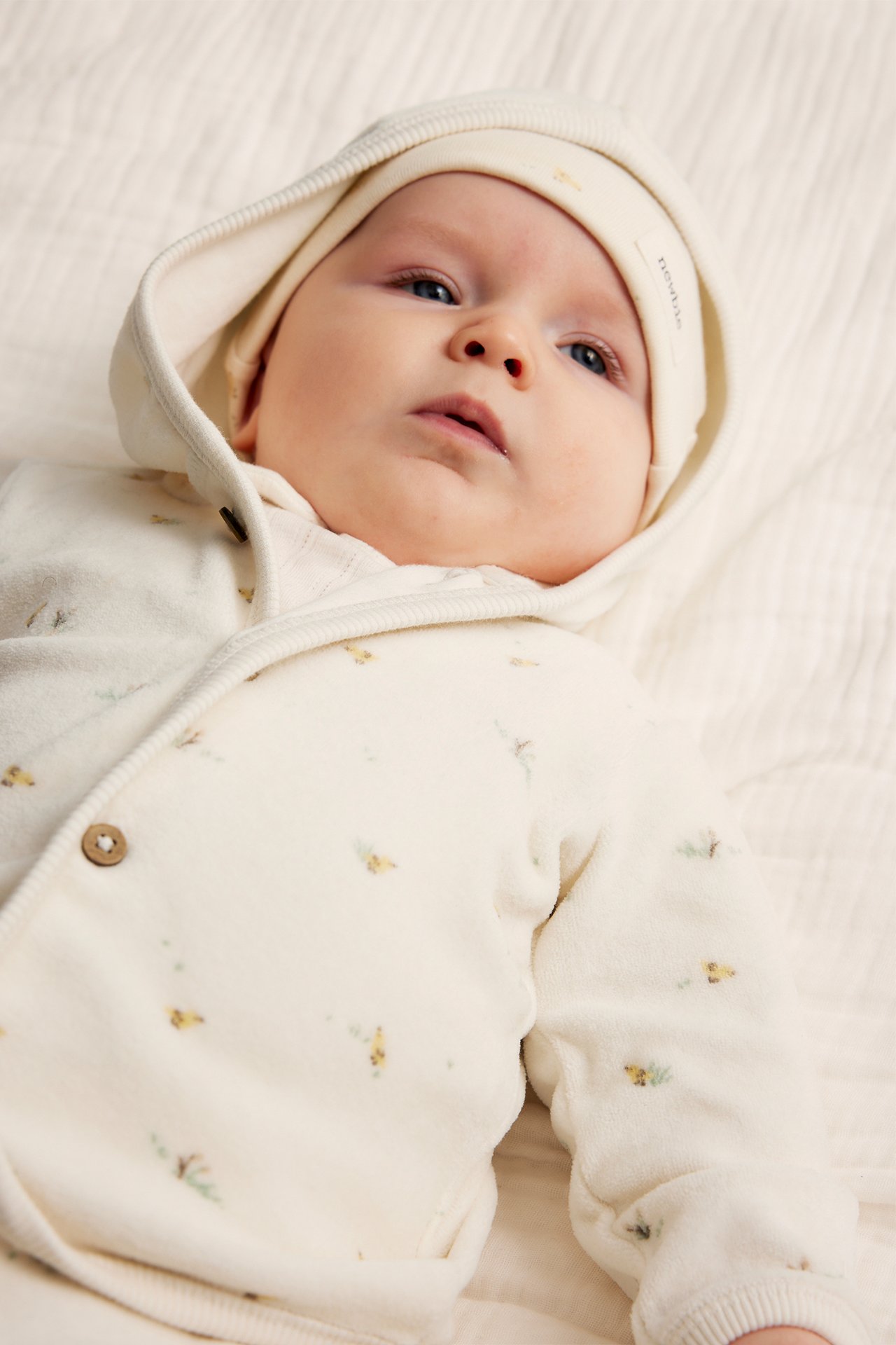 Vorderansicht eines Babys in einem cremefarbenen gemusterten Velours-Kapuzenpullover mit Holzknöpfen und passender Mütze.