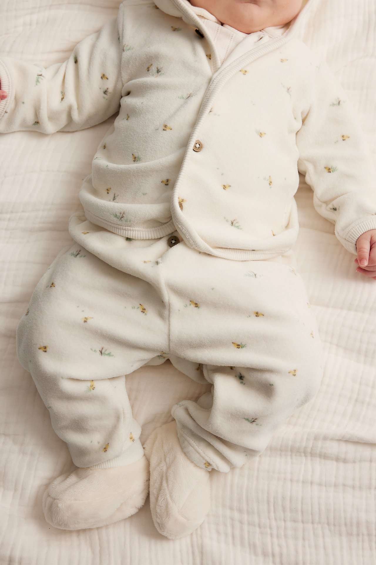 Draufsicht auf Baby in cremeweißem gemustertem Velours-Kapuzenstrickjacke, passender Hose und weichen Schühchen.