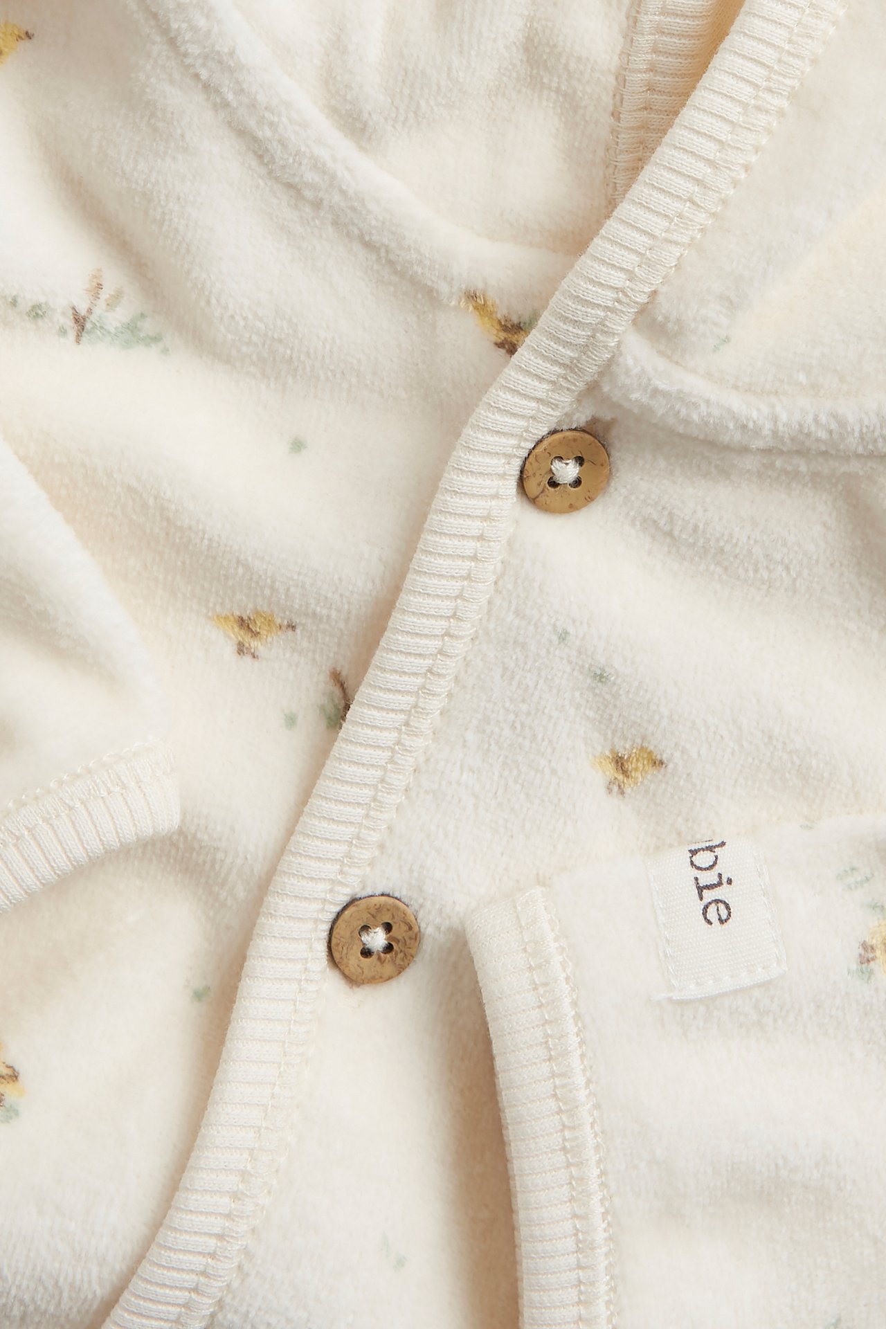 Nahaufnahme eines cremeweißen gemusterten Velours-Baby-Hoodies mit Holzknöpfen vorne.