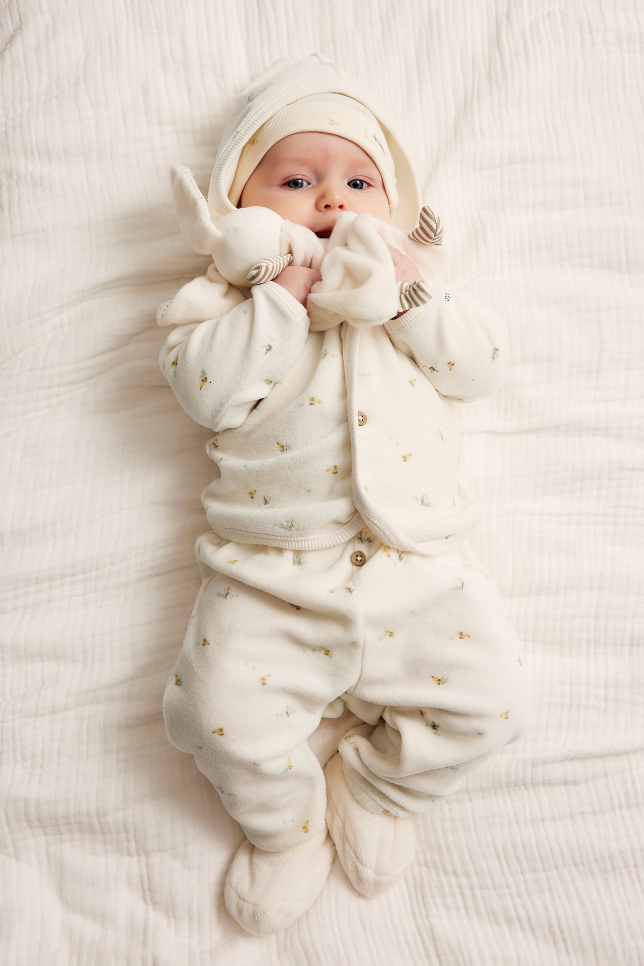 Draufsicht: Baby in cremefarbenem gemustertem Velours-Kapuzenpullover, passender Hose, Mütze, Schühchen, hält Spielzeug.