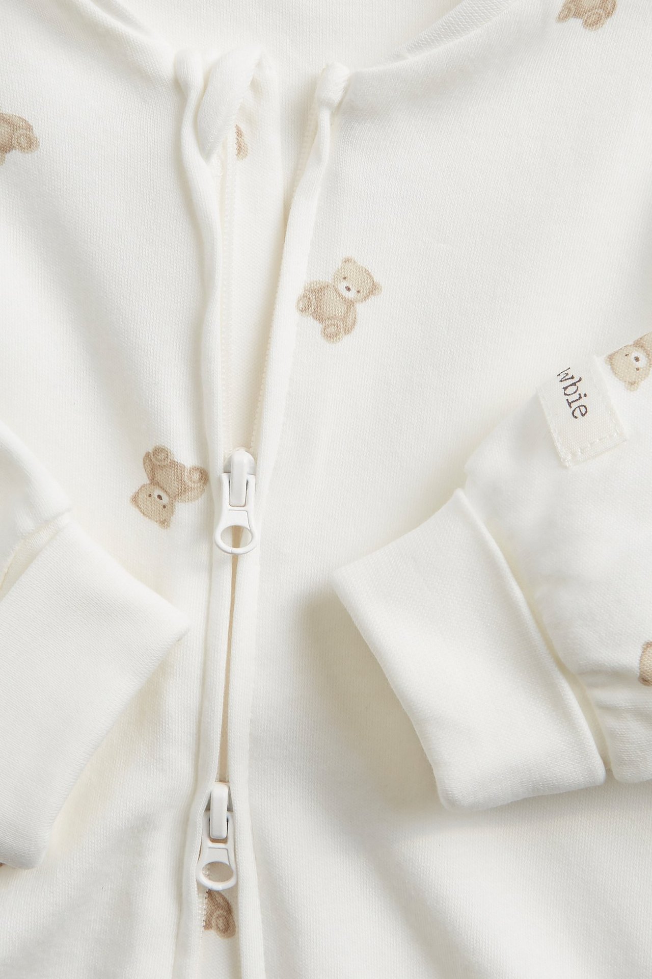 Nahaufnahme eines cremefarbenen Baby-Stramplers aus Baumwolle mit Teddybärmuster, Reißverschluss und umklappbaren Bündchen.