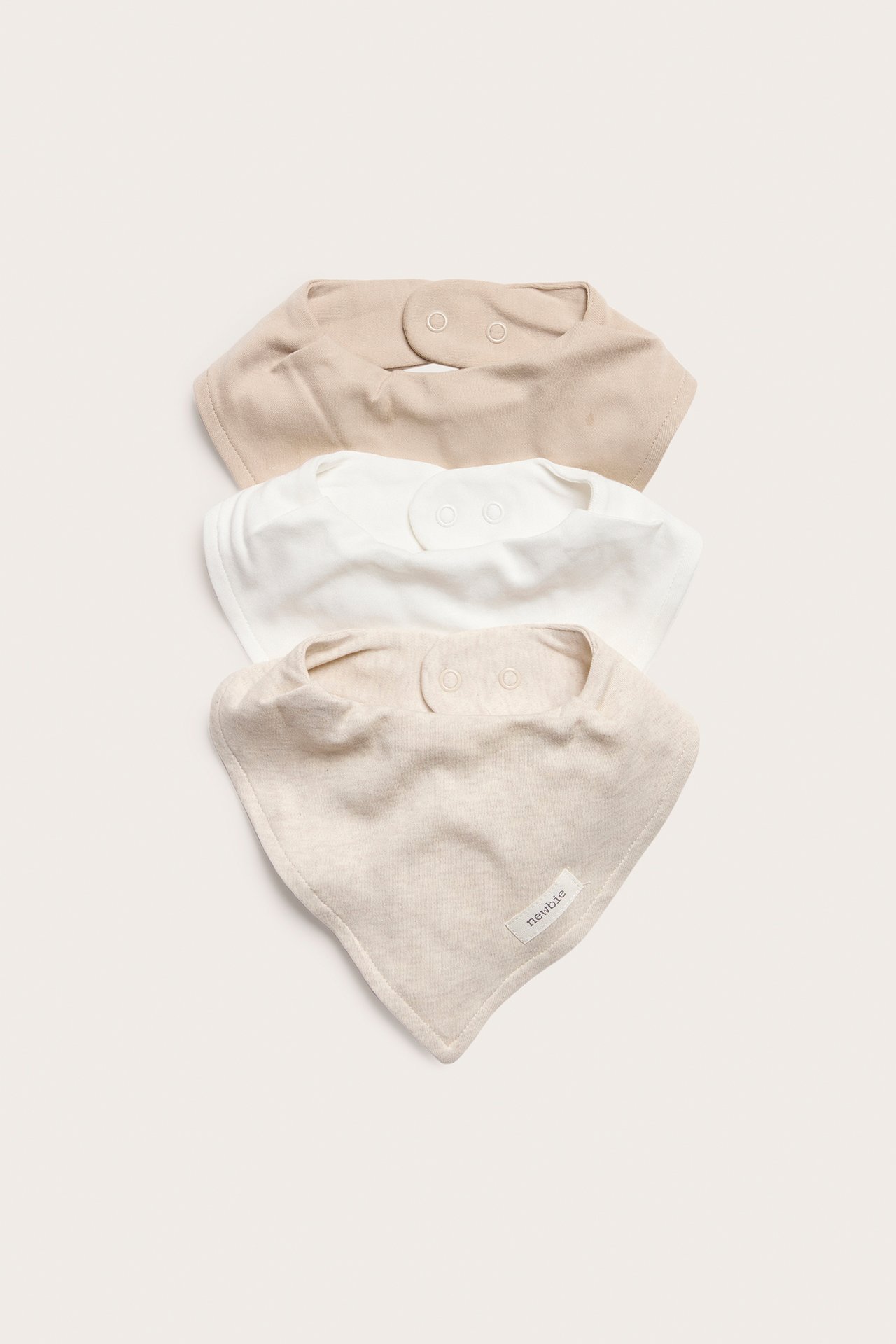 Draufsicht auf drei einfarbige Baby-Lätzchen: Hellbeige, Weiß und Beige Melange, mit Druckknopfverschlüssen.