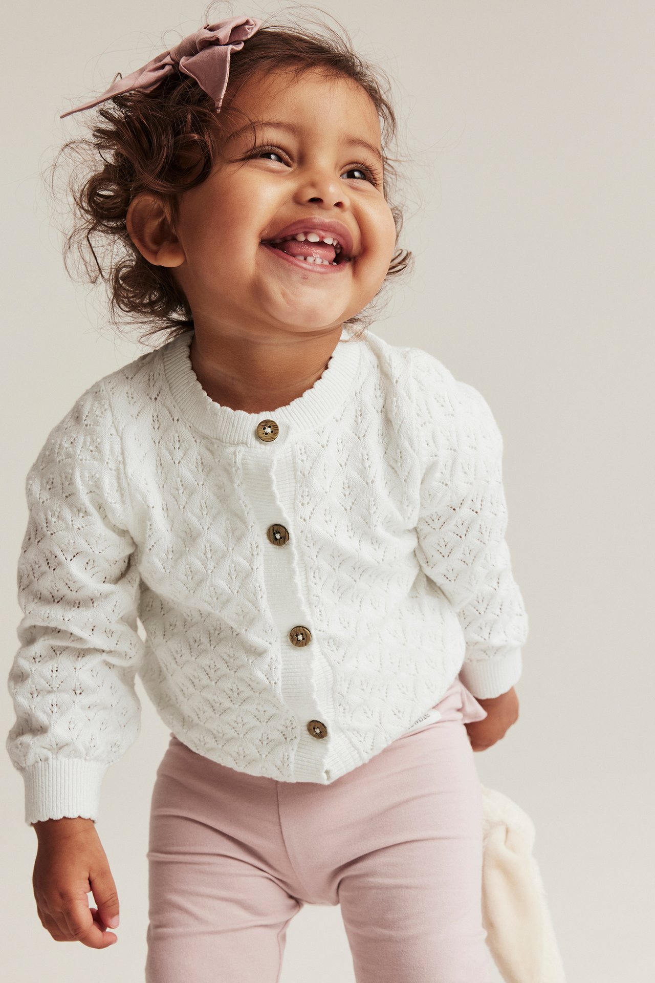 Vorderansicht: Lächelndes Baby in cremeweißer gemusterter Baumwollstrickjacke mit Knöpfen, hellrosa Leggings und rosa Schleife.