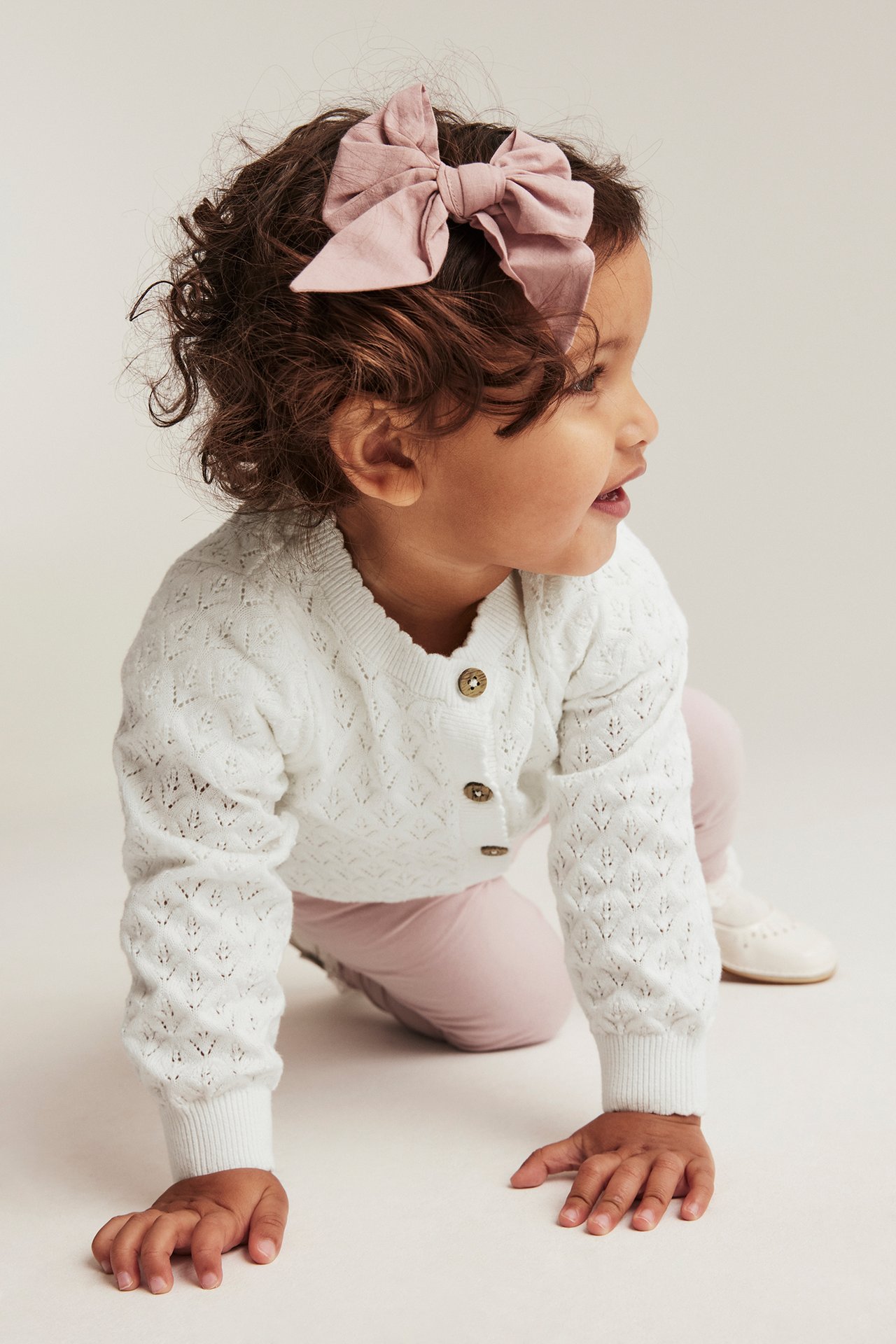 Seitenansicht Baby in cremefarbenem gemustertem Baumwollcardigan mit Knöpfen, hellrosa Hose und rosa Schleife.