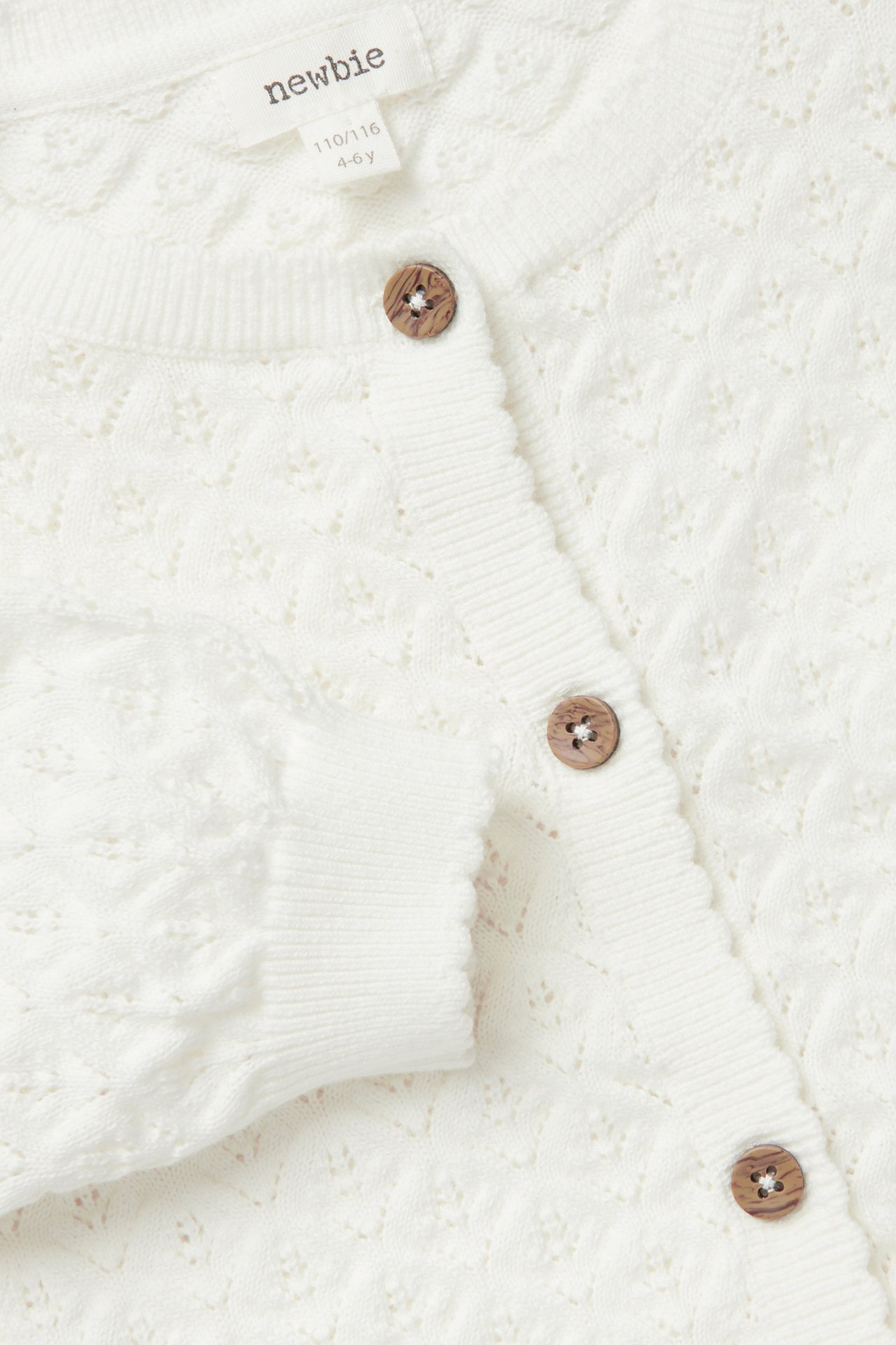 Nahaufnahme Vorderansicht einer cremefarbenen gemusterten Strickjacke für Kinder mit Holzknöpfen und Rippbündchen.