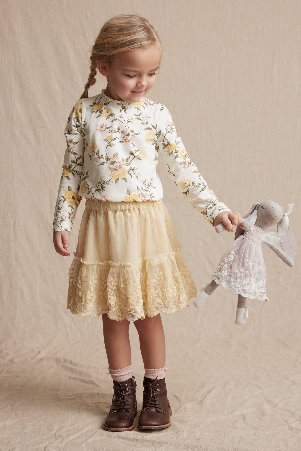 Front view: Kid in offwhite floral jersey top, lace neck, yellow skirt, brown boots, holding doll.