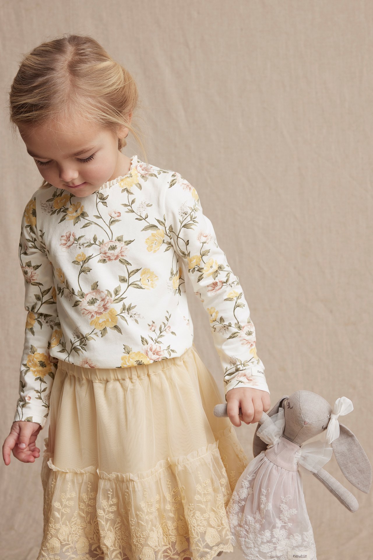 Front view: Kid wears off-white floral jersey top with lace, beige tiered skirt, holding a bunny doll.