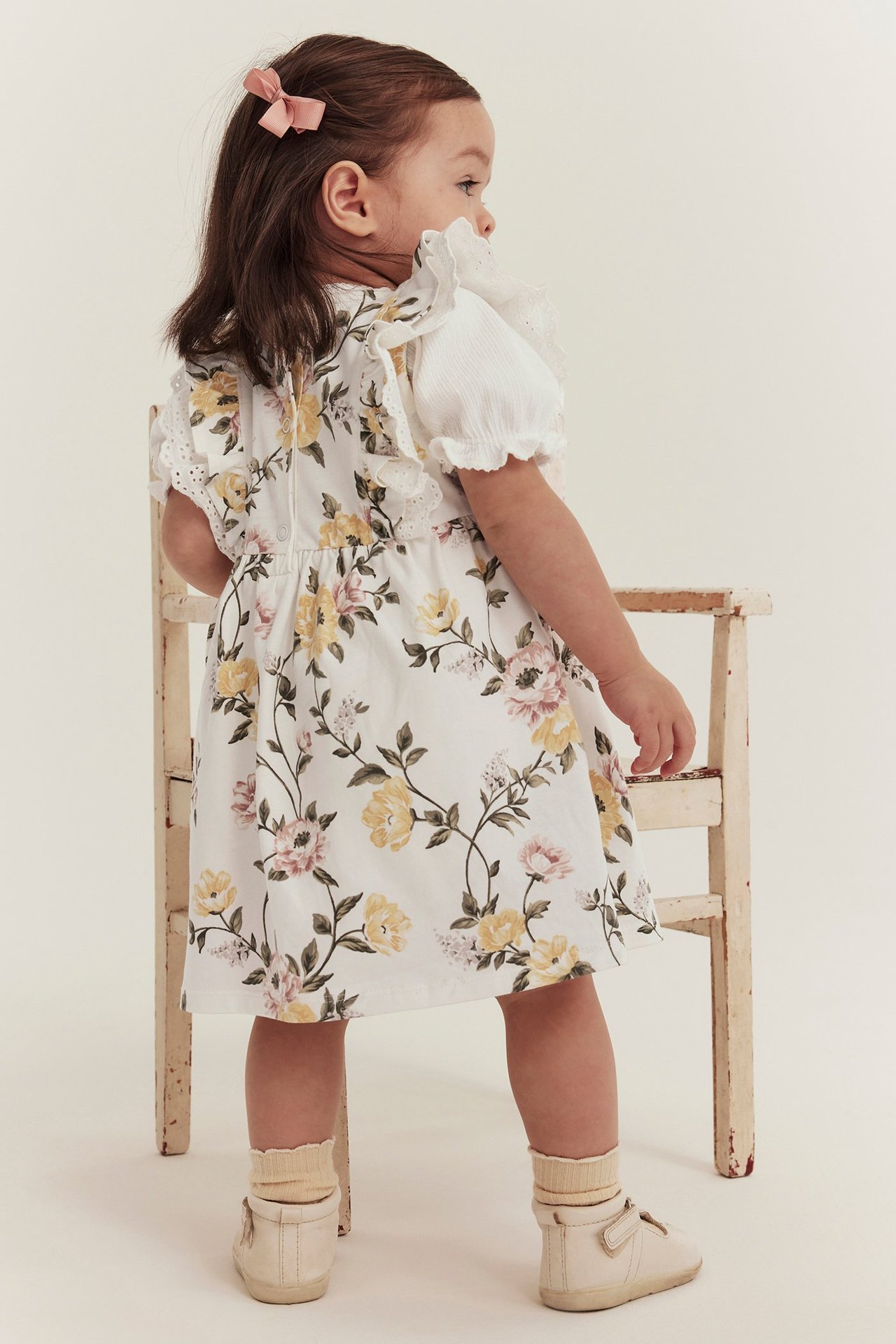 Back view of a kid in an off-white floral dress with white shoulder ruffles, beige socks, and shoes.