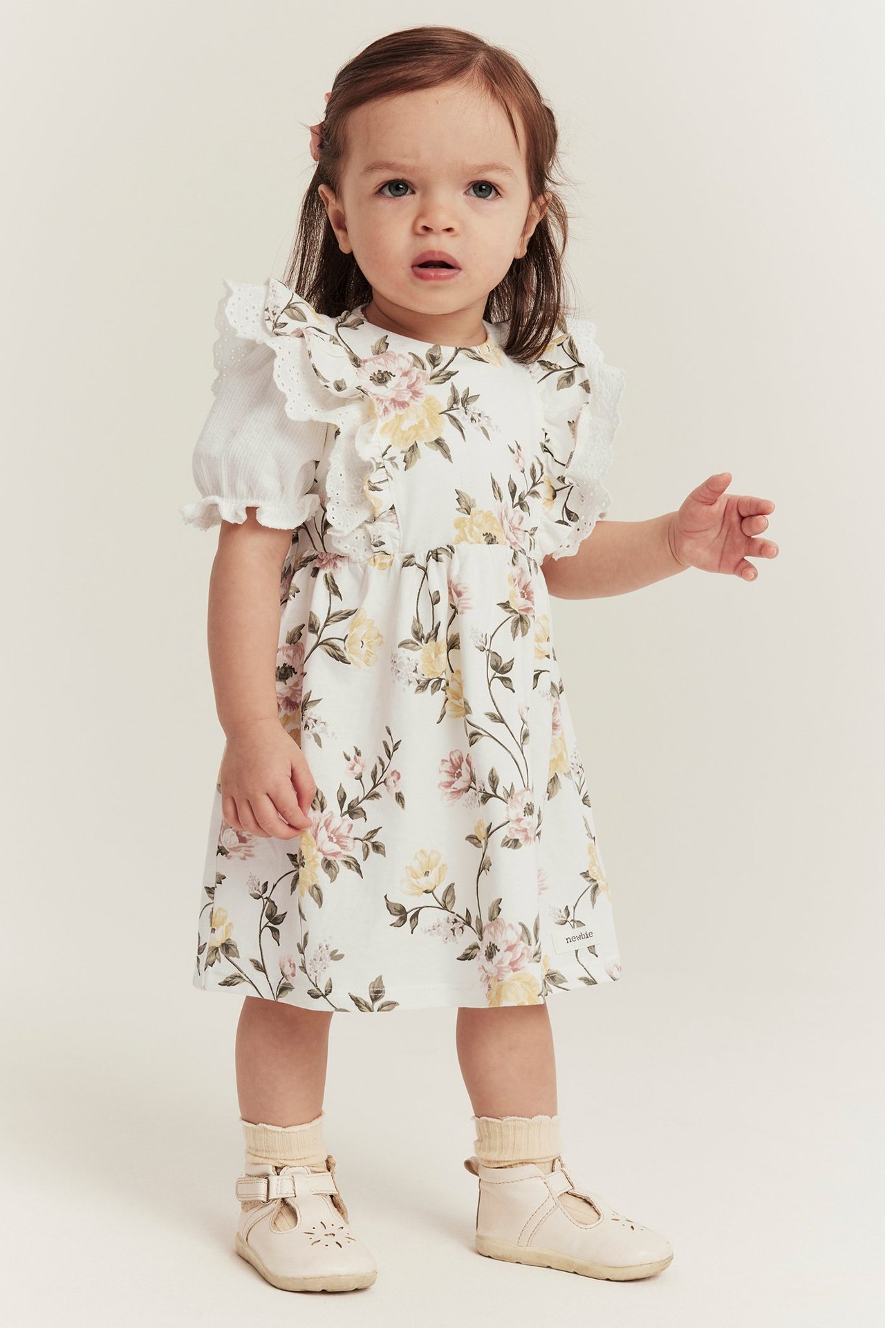 Front view: Baby in off-white floral cotton dress, white ruffled blouse, beige socks, and T-bar shoes.