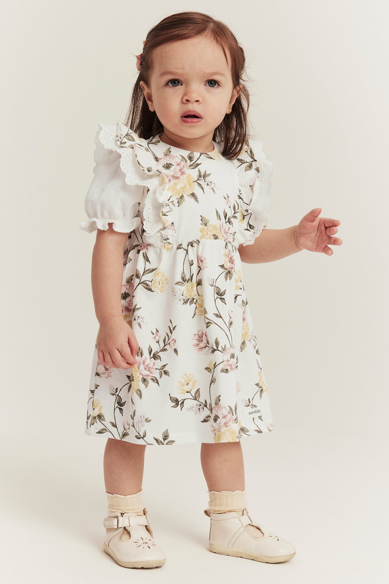 Vorderansicht: Baby in cremeweißem Blumenkleid, weißer Rüschenbluse, beigen Socken und T-Spangen-Schuhen.