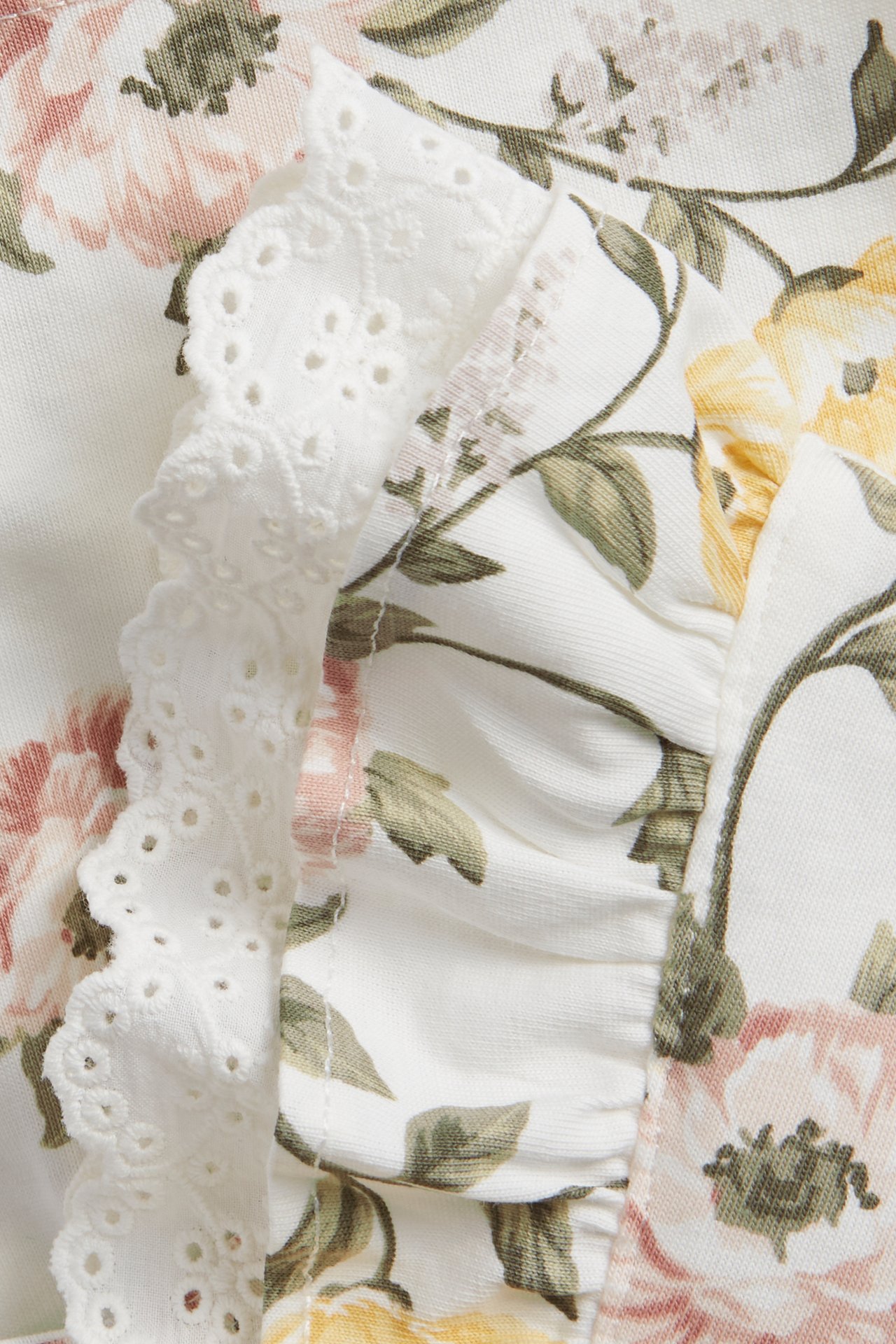 Nahaufnahme eines cremefarbenen Baumwollkleides mit Blumenmuster, rosa und gelben Blüten, mit weißer Spitze und Stoffrüsche.