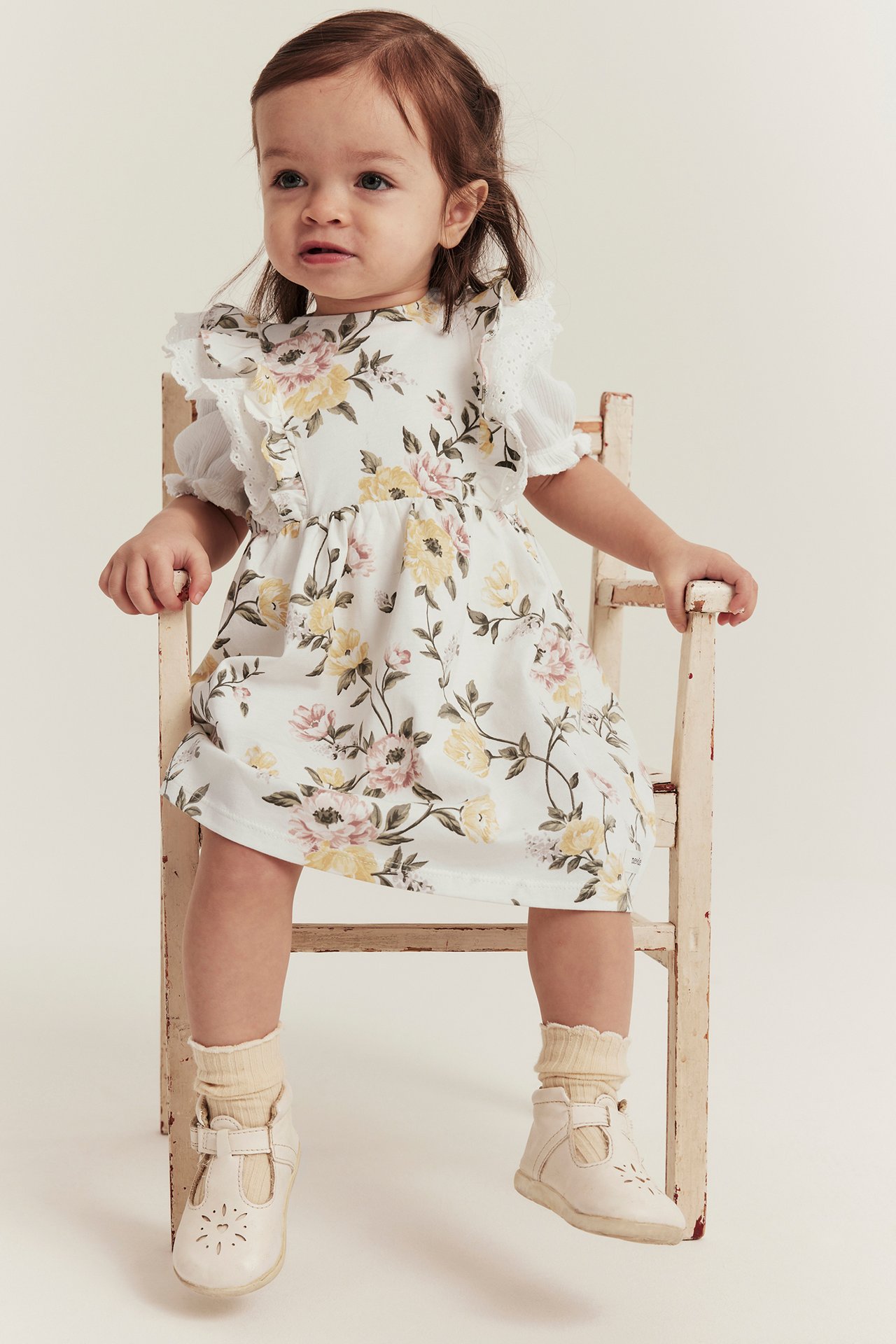 Vorderansicht: Baby in cremeweißem Blumenkleid mit Rüschen, weißem Oberteil, cremefarbenen Socken und T-Spangen-Schuhen.