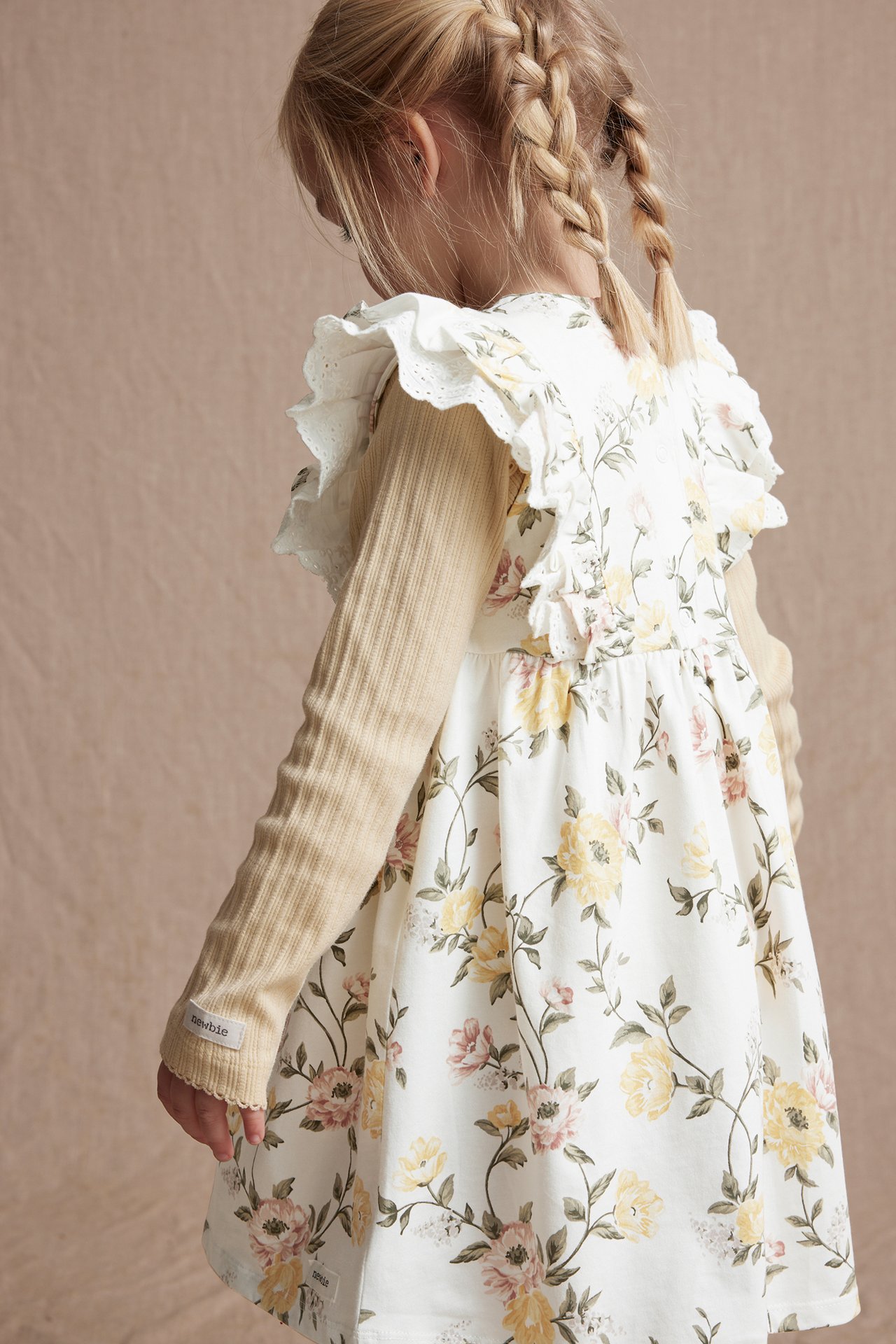 Kind, Seitenansicht von hinten: Off-weißes Blumenkleid mit Rüschen, getragen über einem beigen gerippten Langarmshirt.