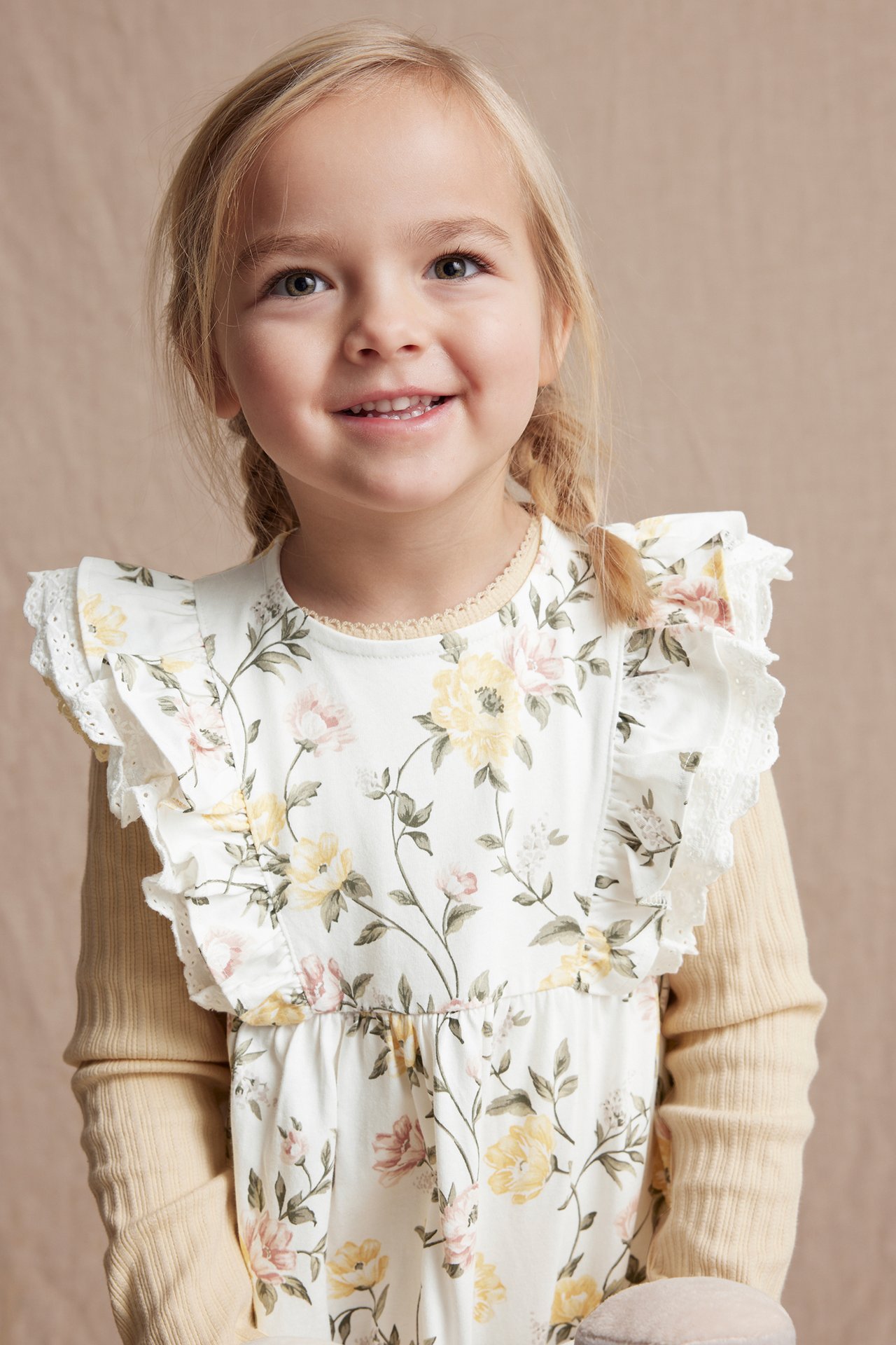 Vorderansicht: Kind in einem cremefarbenen geblümten Baumwollkleid mit Spitzenrüschen über einem hellbeigen Top.